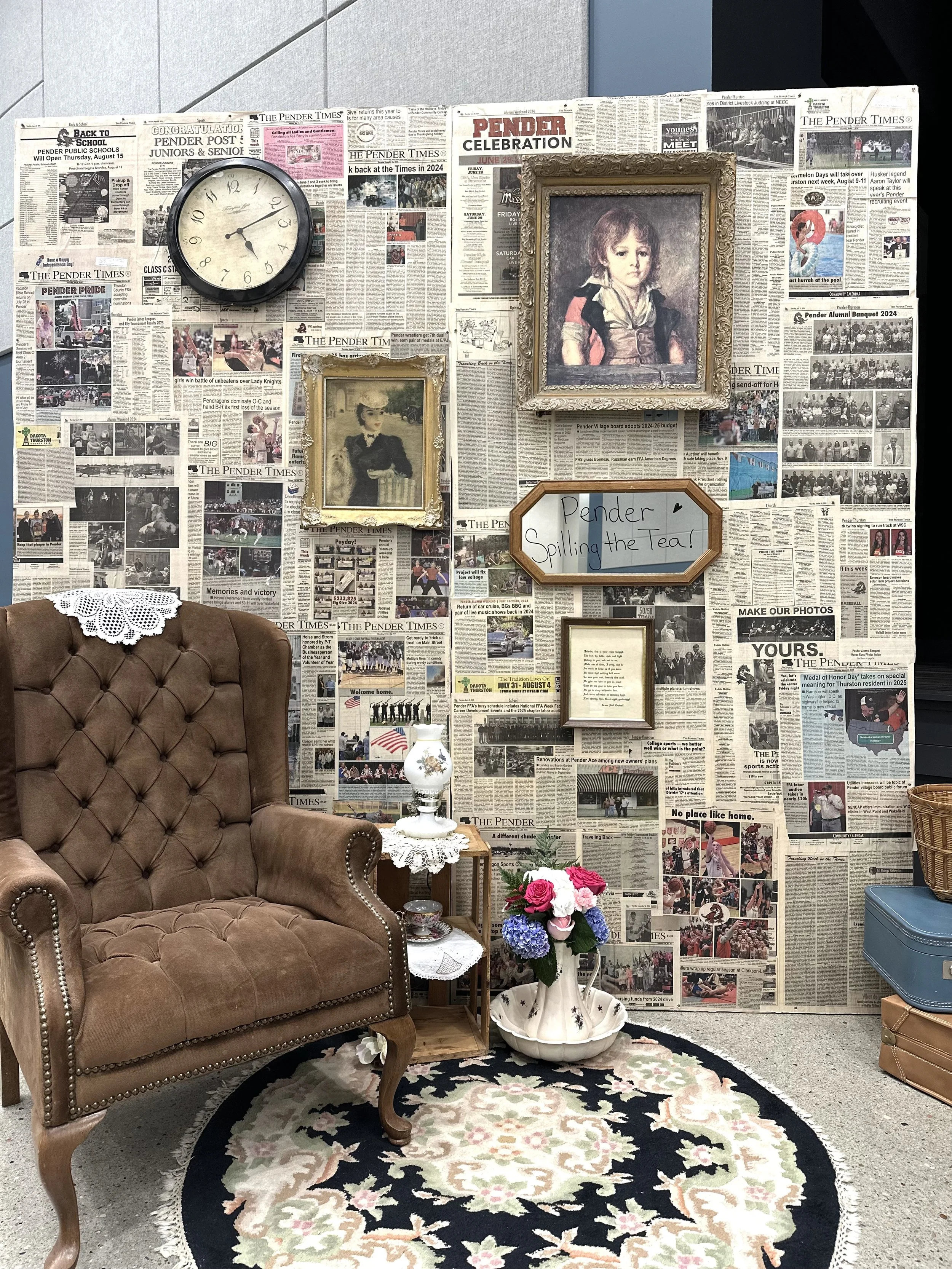 A vintage-style room with a brown tufted armchair, a small wooden side table with a lace doily, a floral arrangement, and a patterned rug. The wall behind is covered with newspapers, decorated with framed photos, a whiteboard with handwritten text, a