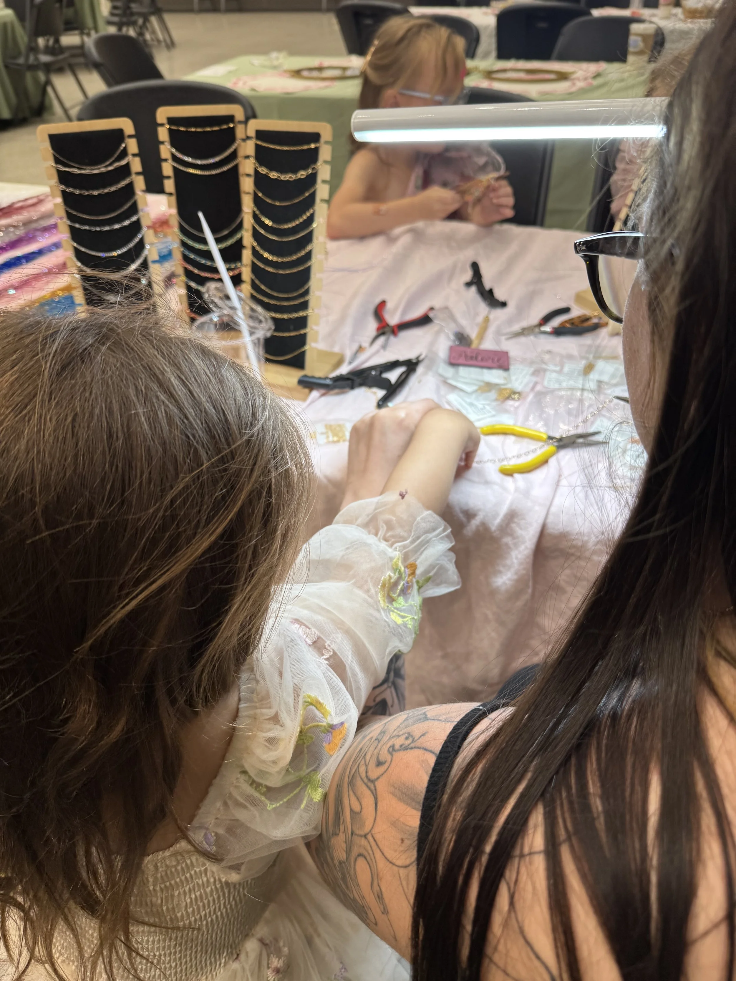 A woman and a young girl working on jewelry at a craft table, with jewelry tools, pliers, and display stands with necklaces and earrings.