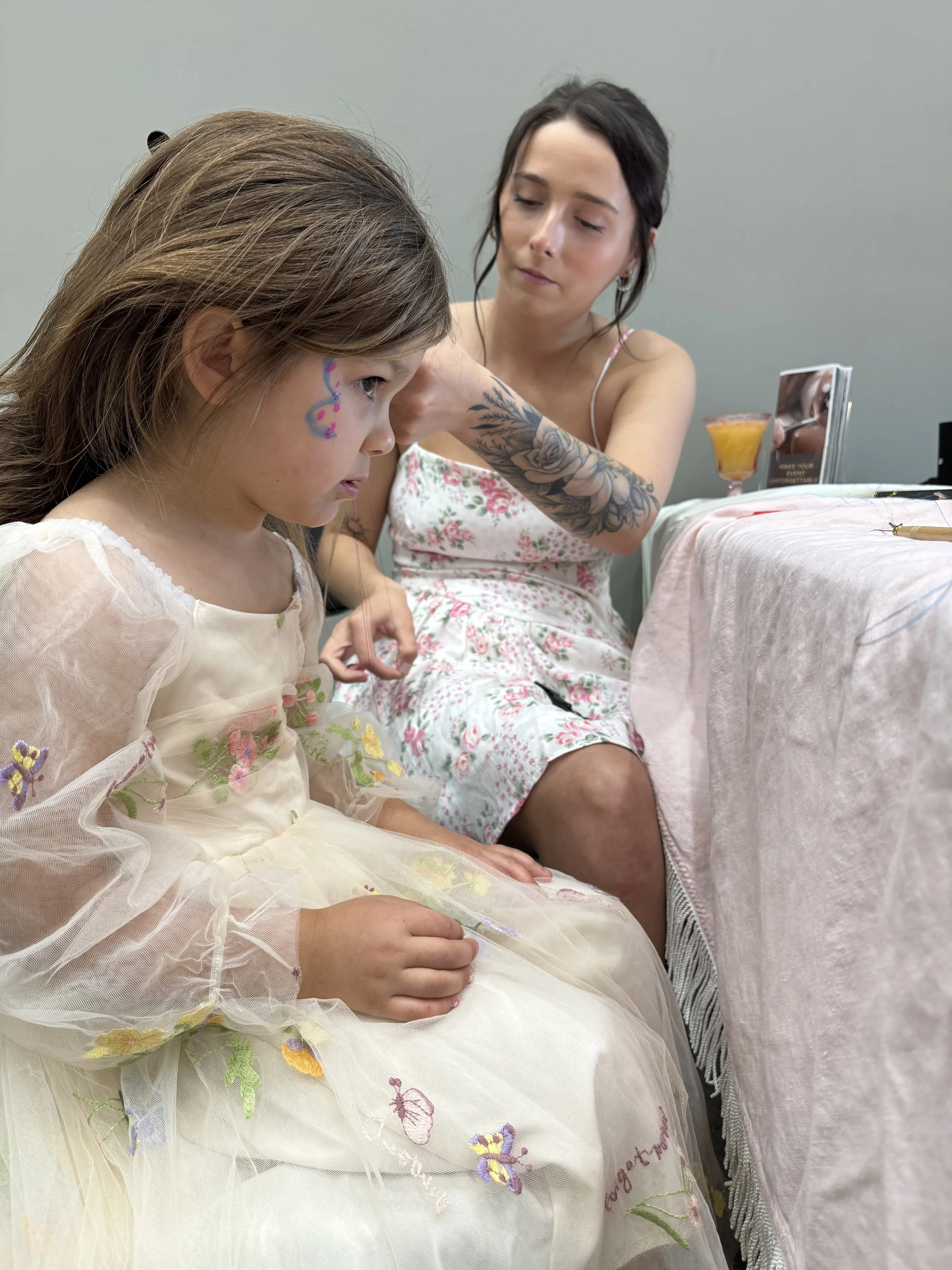 A young girl in a light-colored dress with floral embroidery and face paint sits at a table. An adult woman with dark hair and a tattooed arm is leaning over her, appearing to help or decorate her. The table has a soft pink cover, and there are drink