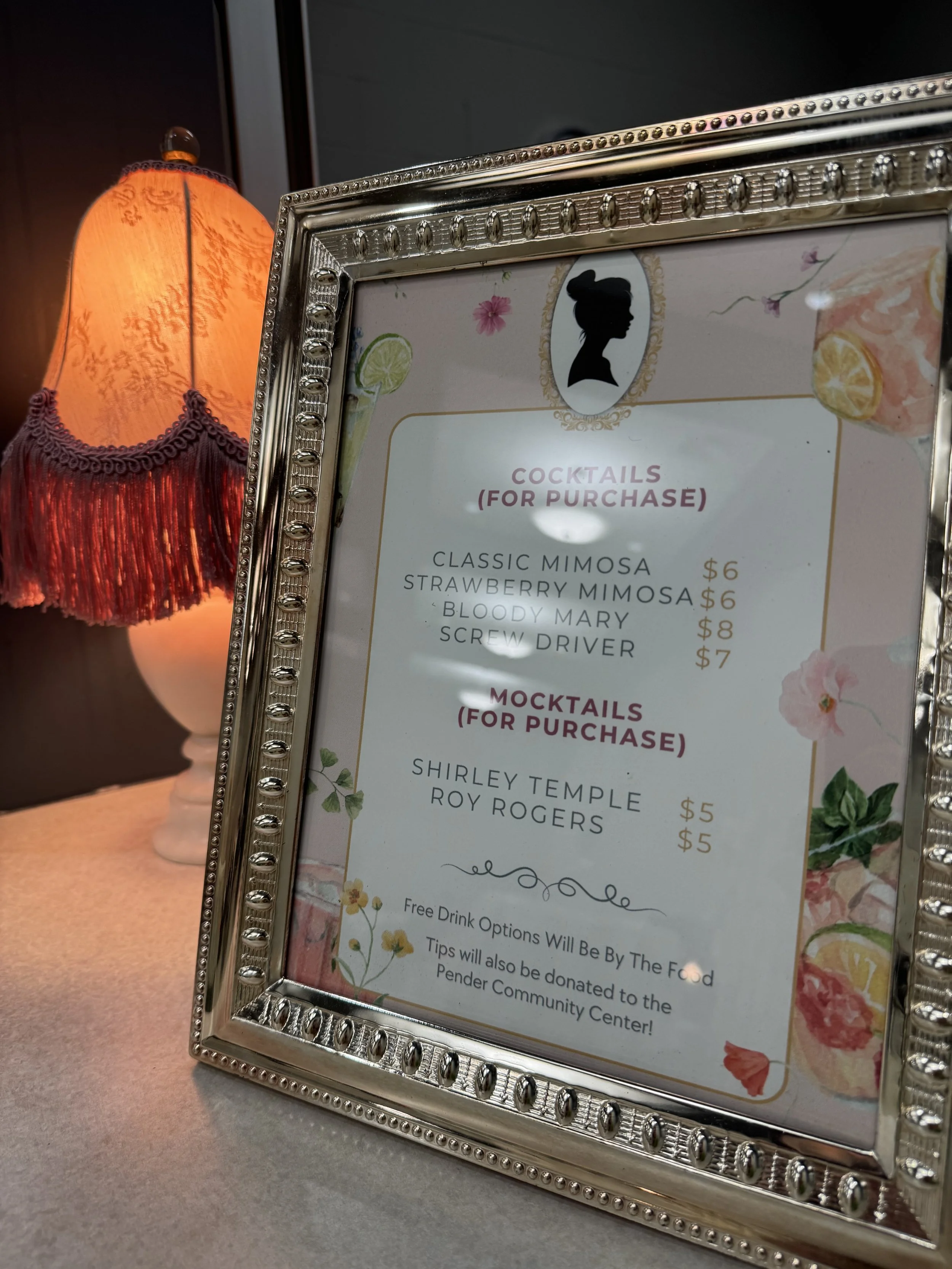 Framed menu listing cocktails and mocktails with prices, next to a lamp with an orange shade and red fringe.