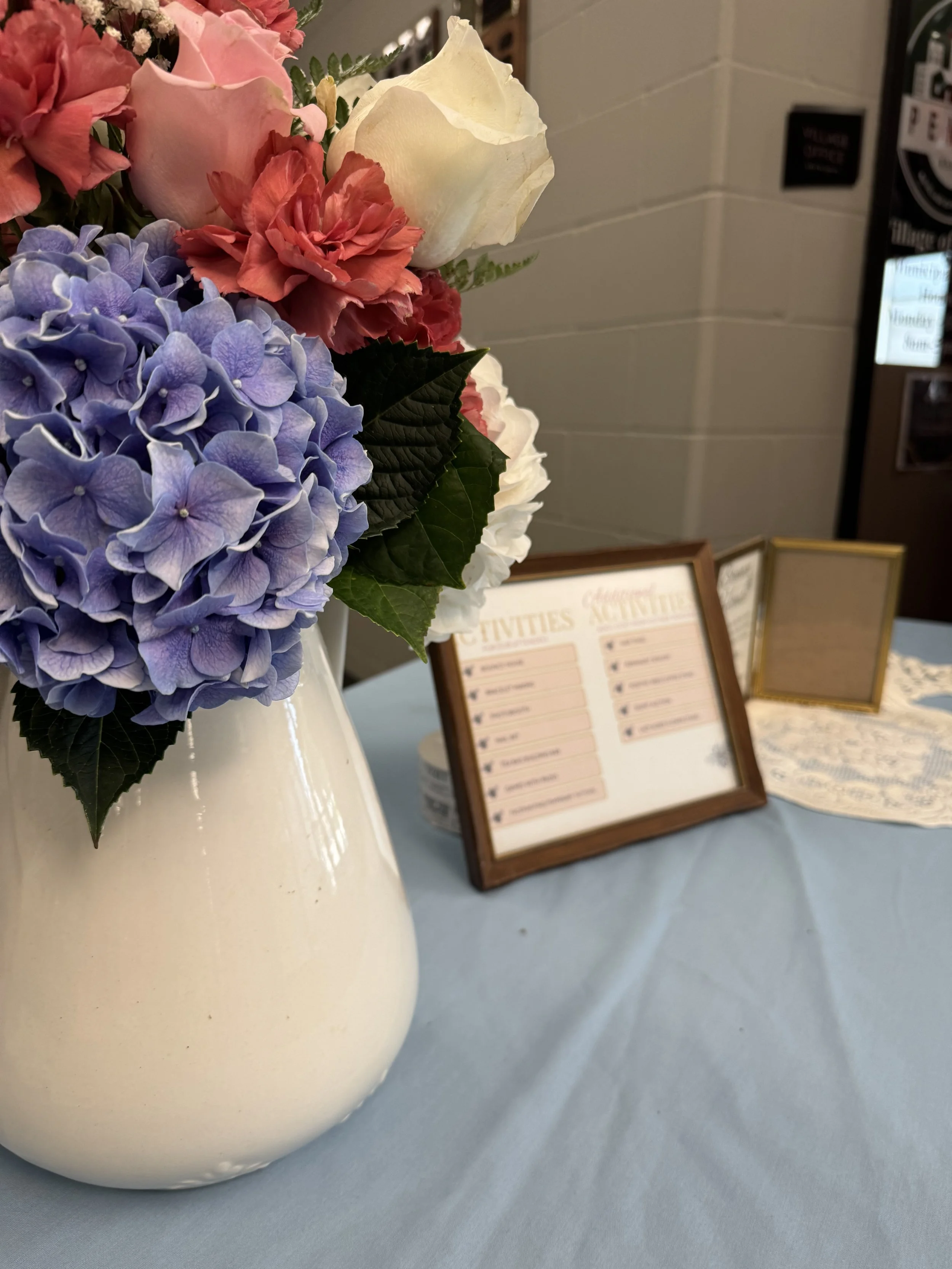 Close-up of a floral arrangement with purple hydrangeas, pink roses, white roses, and green leaves in a white vase on a light blue tablecloth. In the background, there are framed signs or menus and a white doily.