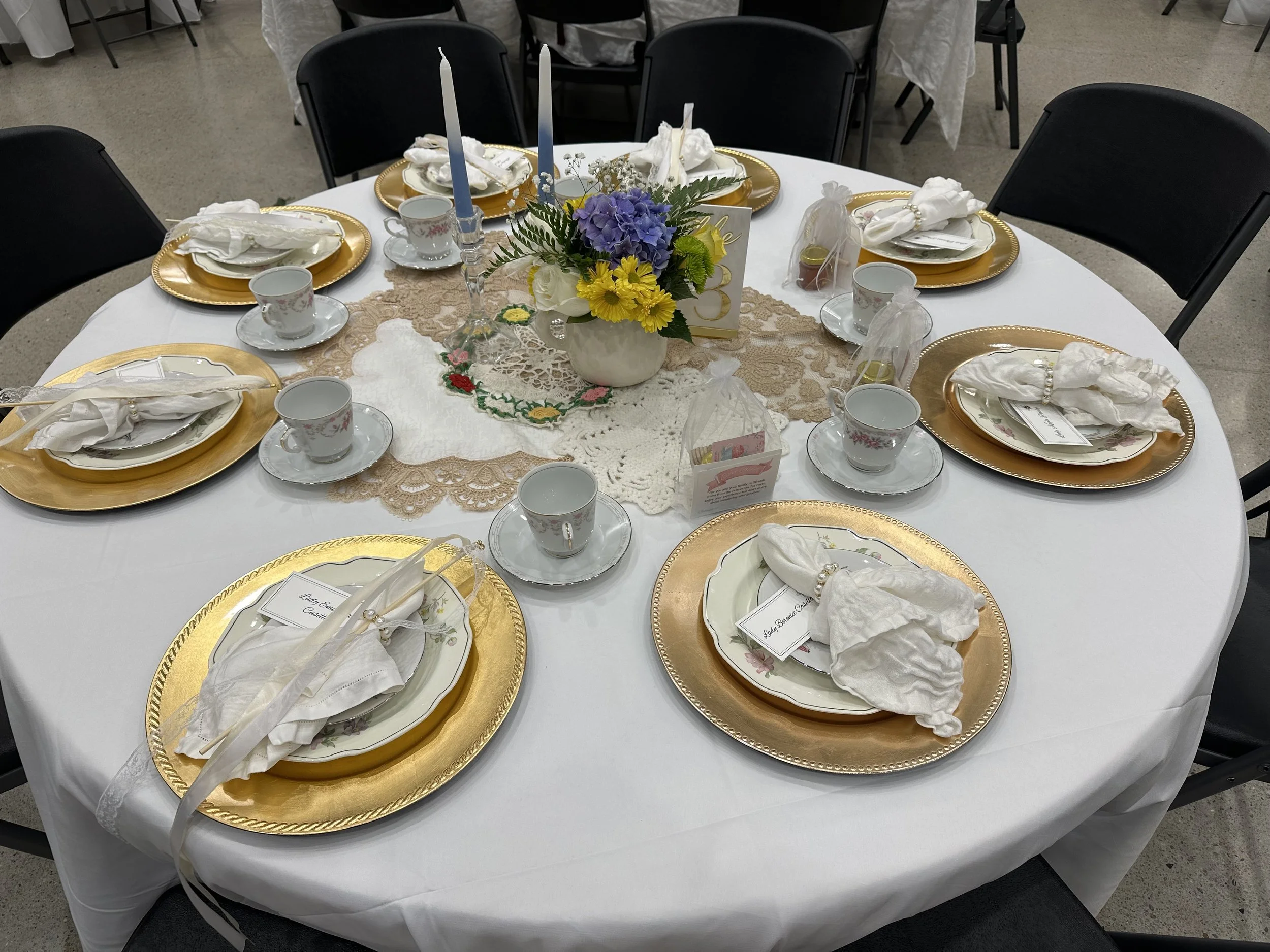 Round table set for a formal occasion with white tablecloth, gold chargers, china plates, teacups, cloth napkins, and a central floral arrangement of purple, yellow, and white flowers. Place cards and small favor bags are also on the table.