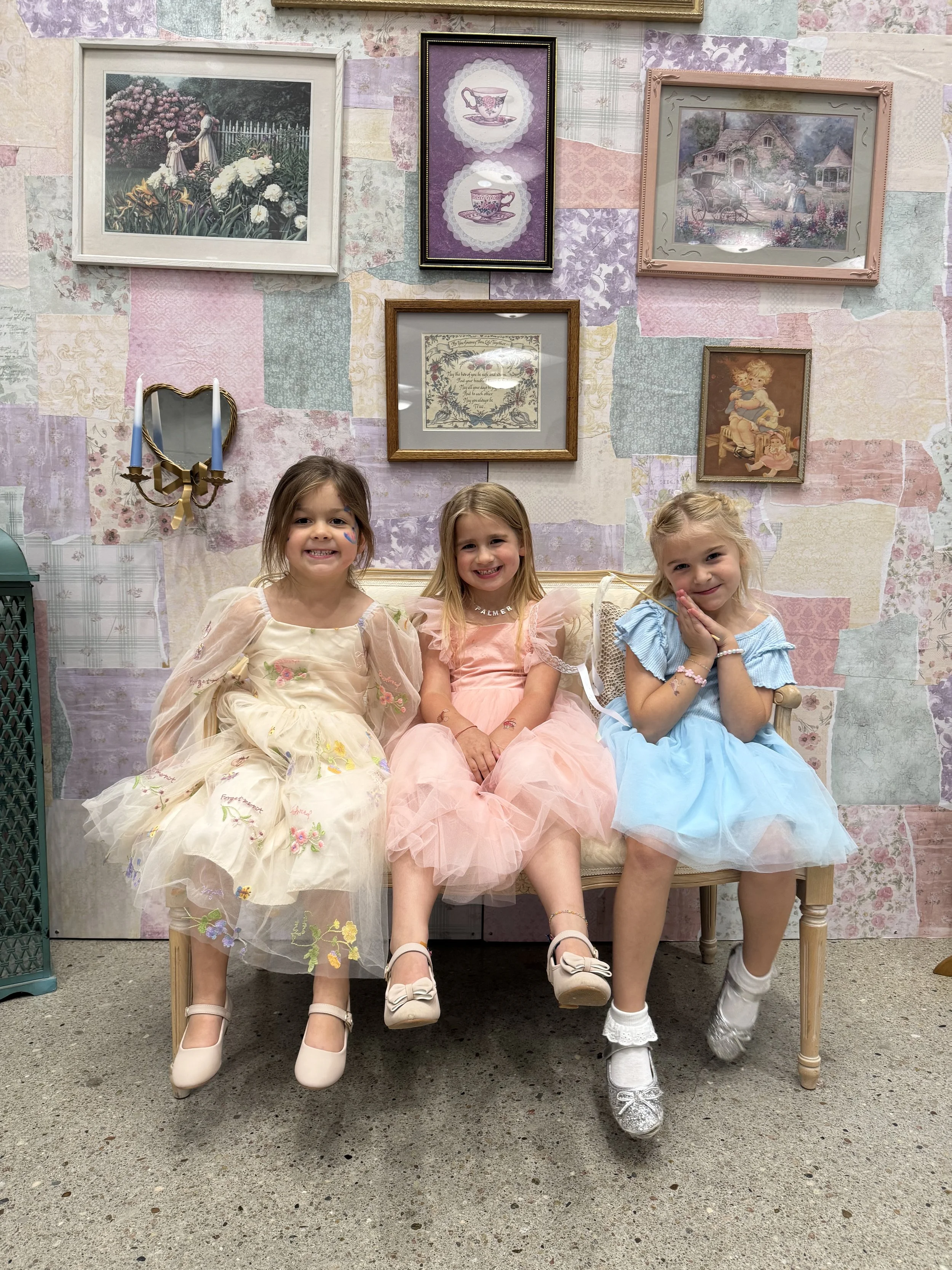 Three young girls in pastel-colored dresses sitting on a vintage-style bench, smiling at the camera. The background features a decorated wall with framed artwork and floral patterns.