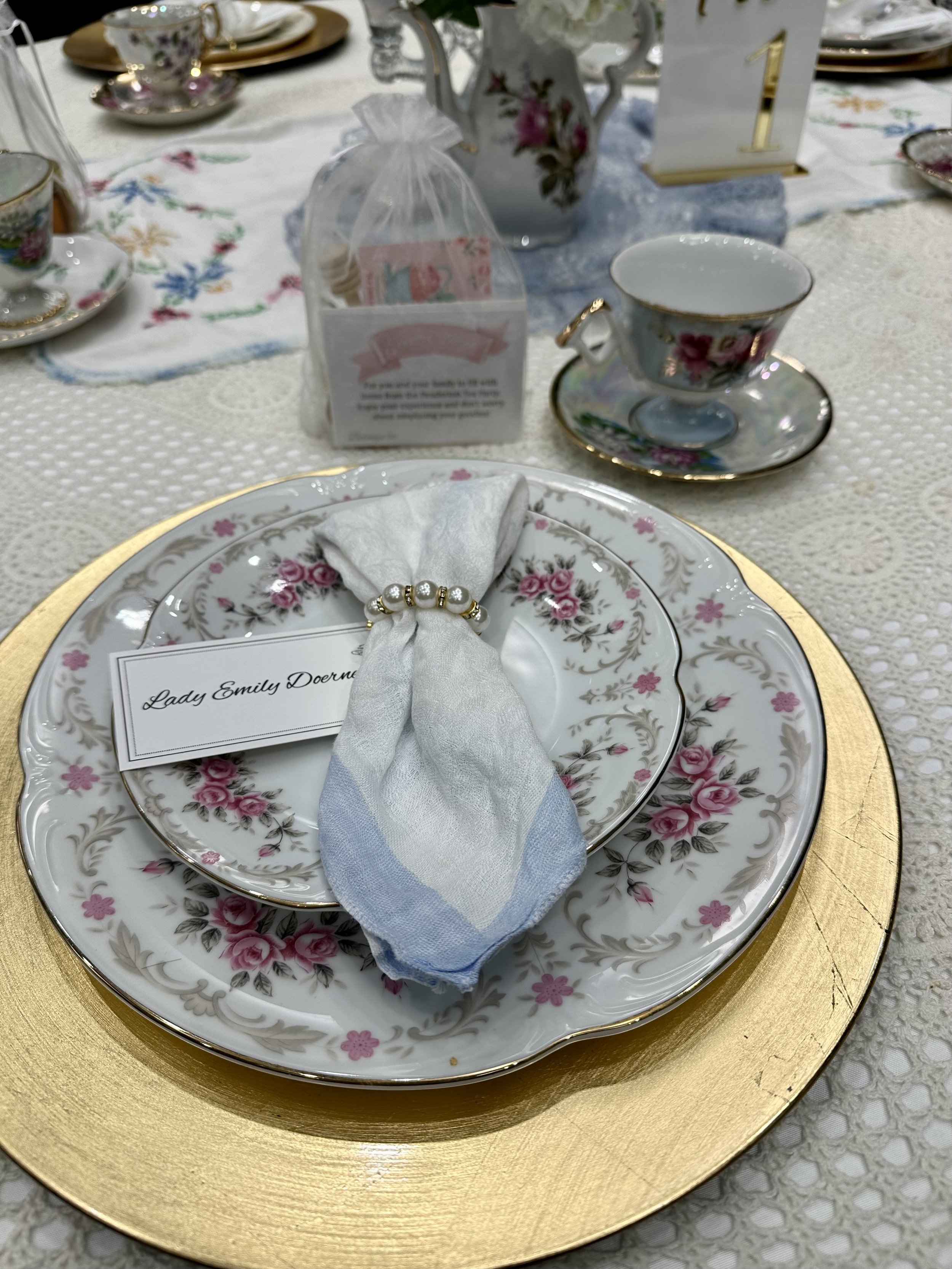 Elegant table setting with floral china plates, a napkin with a pearl napkin ring, a name card for Lady Emily Doerne, fine teacups and saucers, a decorative teapot with flowers, and a gold placemat.