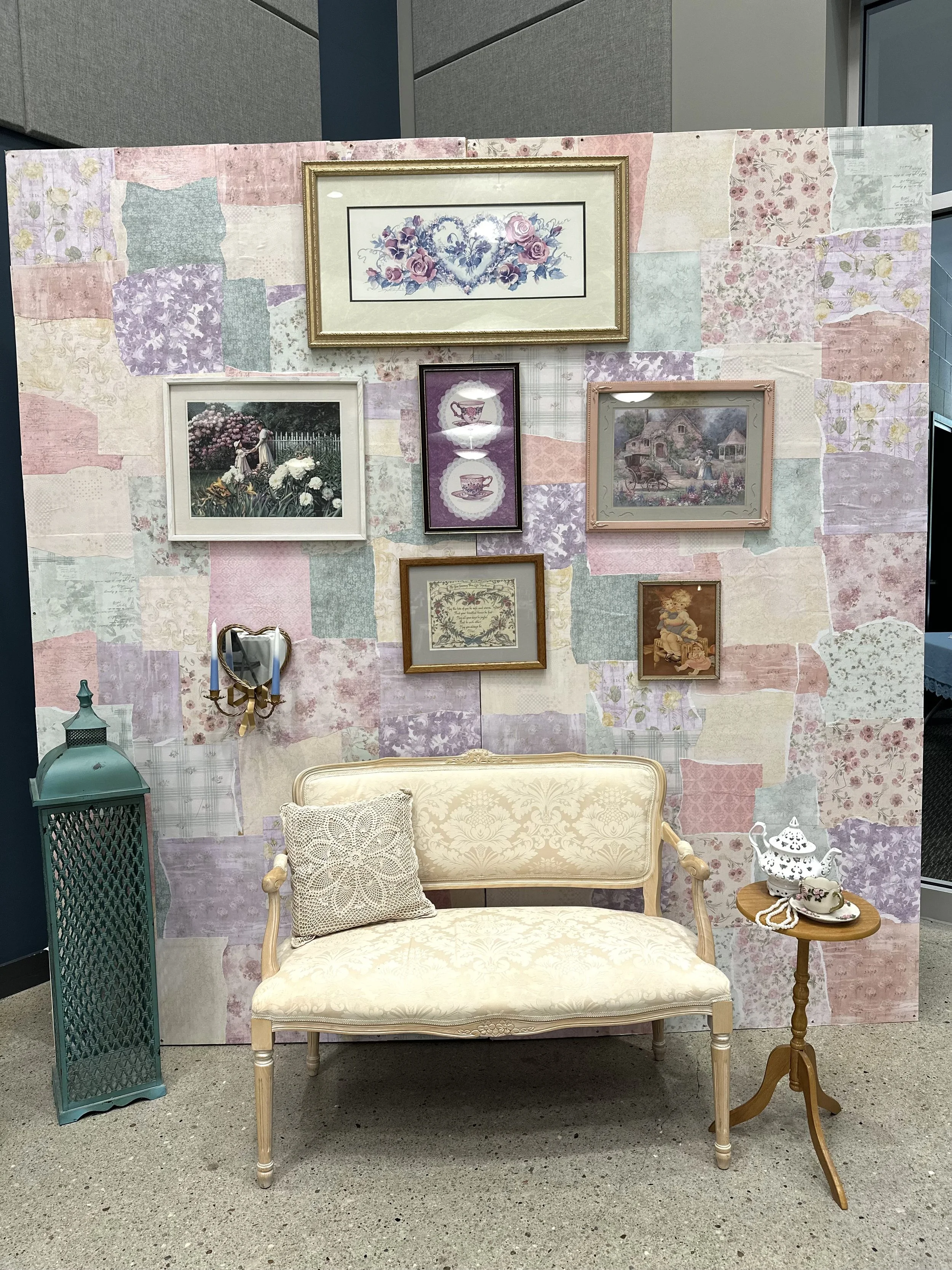 A vintage-style sitting area with a beige upholstered loveseat, decorated with a lace cushion, a patchwork floral fabric backdrop with framed artwork, a teal lantern on the floor, a wooden side table with a white teapot and cup, and a small heart-sha