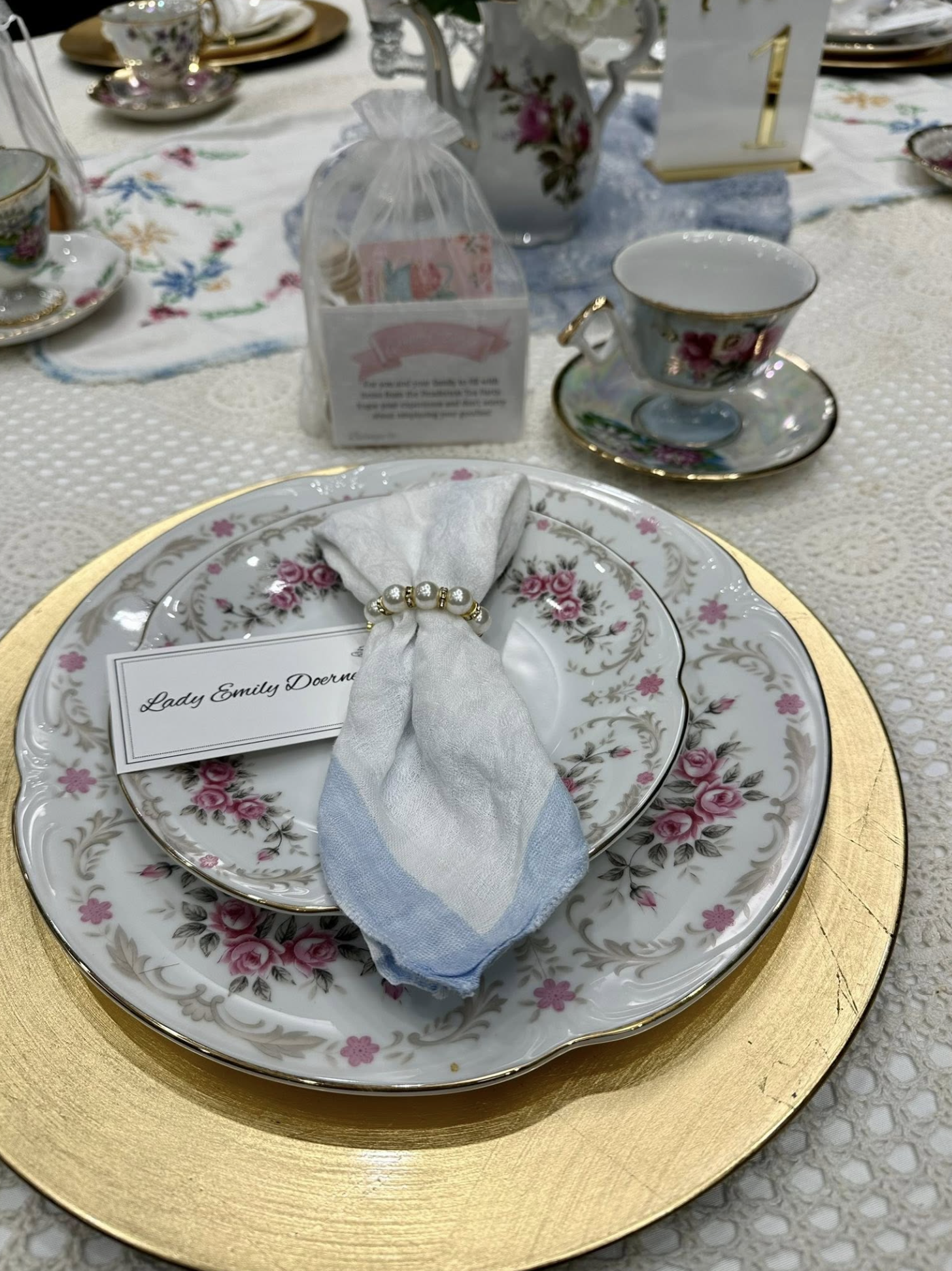 Elegant table setting with floral porcelain plates, a gold charger, a wrapped utensil, and vintage china cups and saucers, decorated for a formal event.