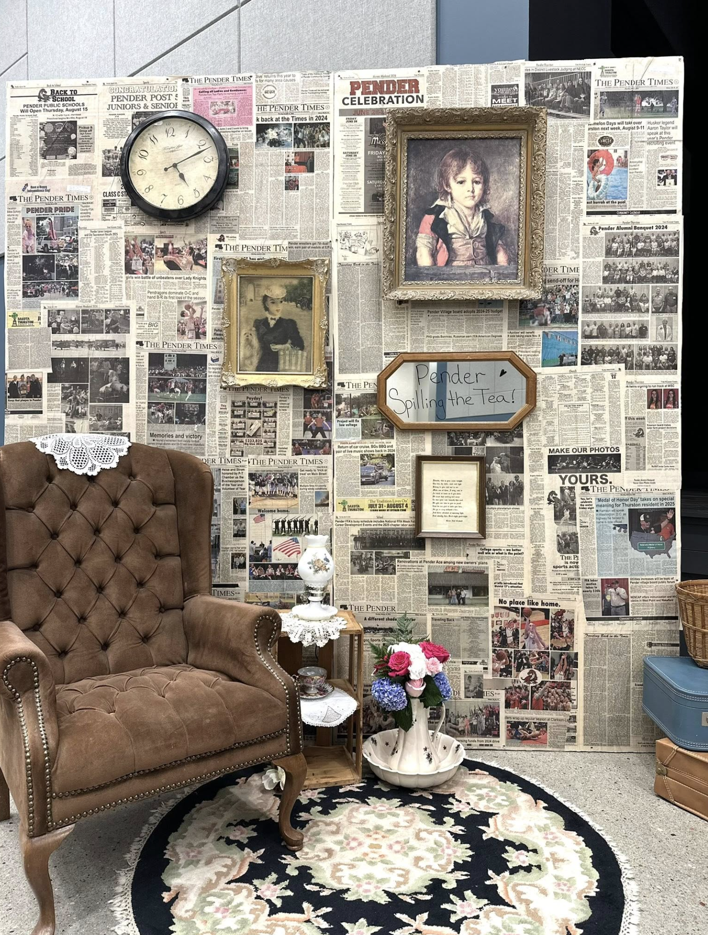 A cozy vintage room with a wall covered in newspapers, a brown tufted armchair, a small wooden side table with a lamp, a flower vase, and a decorative rug. The wall features framed artwork, a clock, and a bulletin board with the note "Pender Spilling the Tea!".
