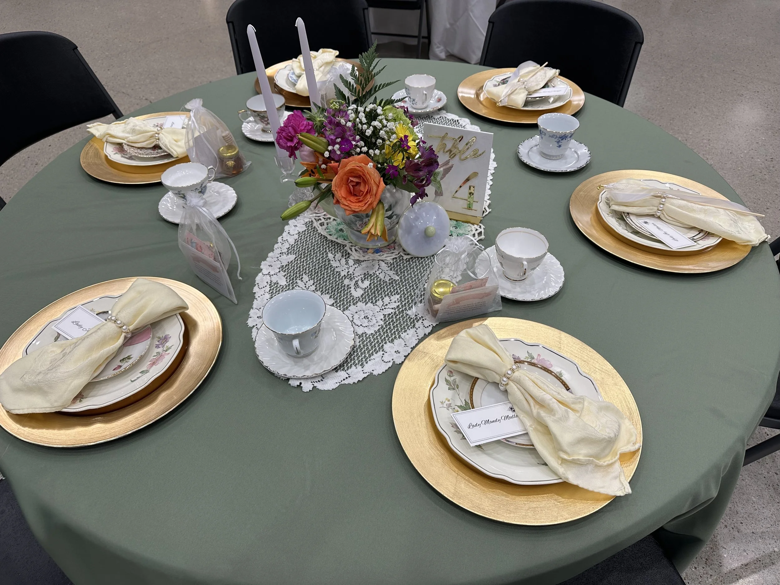 A round table set for a celebration with six place settings, a floral centerpiece with pink, orange, purple, and white flowers, bamboo plates, china plates with floral designs, yellow cloth napkins with pearl napkin rings, teacups and saucers, white 