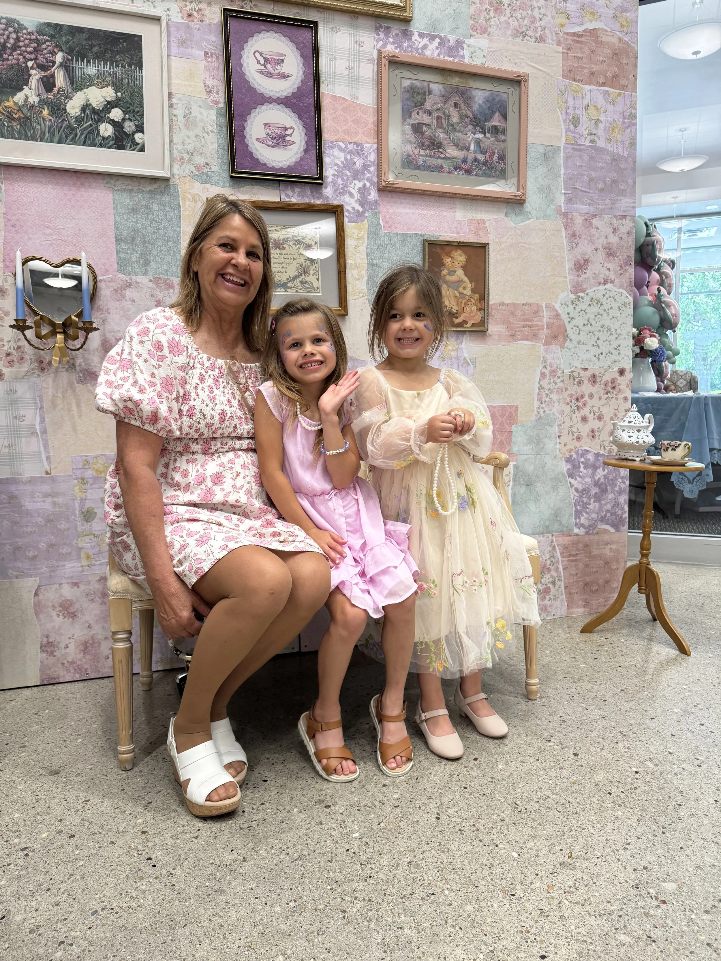 Three women, including a woman and two young girls, sitting on a vintage-style bench at a tea party. They are surrounded by pastel-colored wall art, including floral and tea-themed pictures. The women are dressed in vintage-style dresses, and one gir