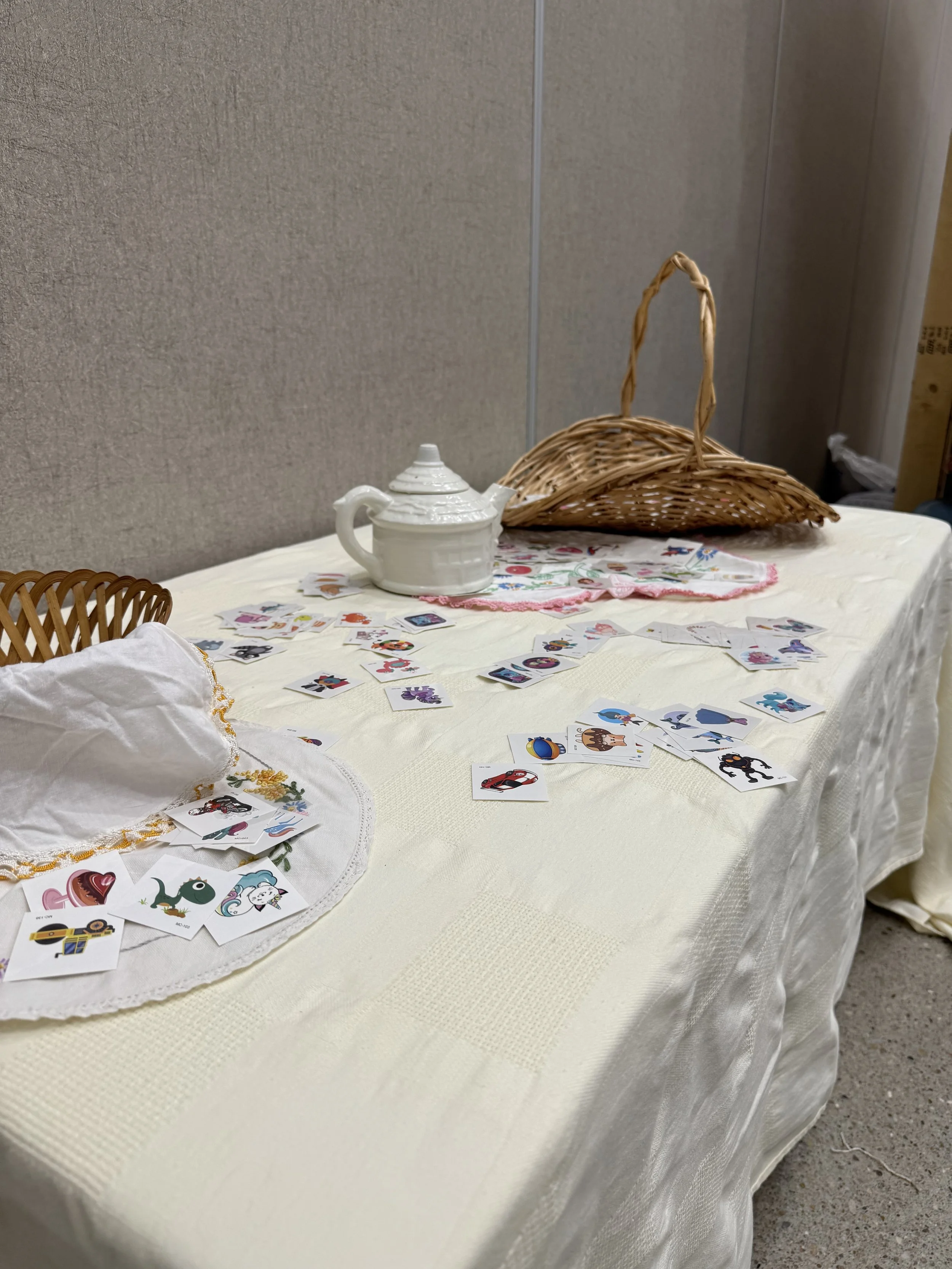 A table covered with a cream-colored cloth, scattered with picture cards featuring various colorful characters and objects, possibly for a children's game or activity. There is a white teapot, a decorative white cloth or napkin, and a wicker basket o