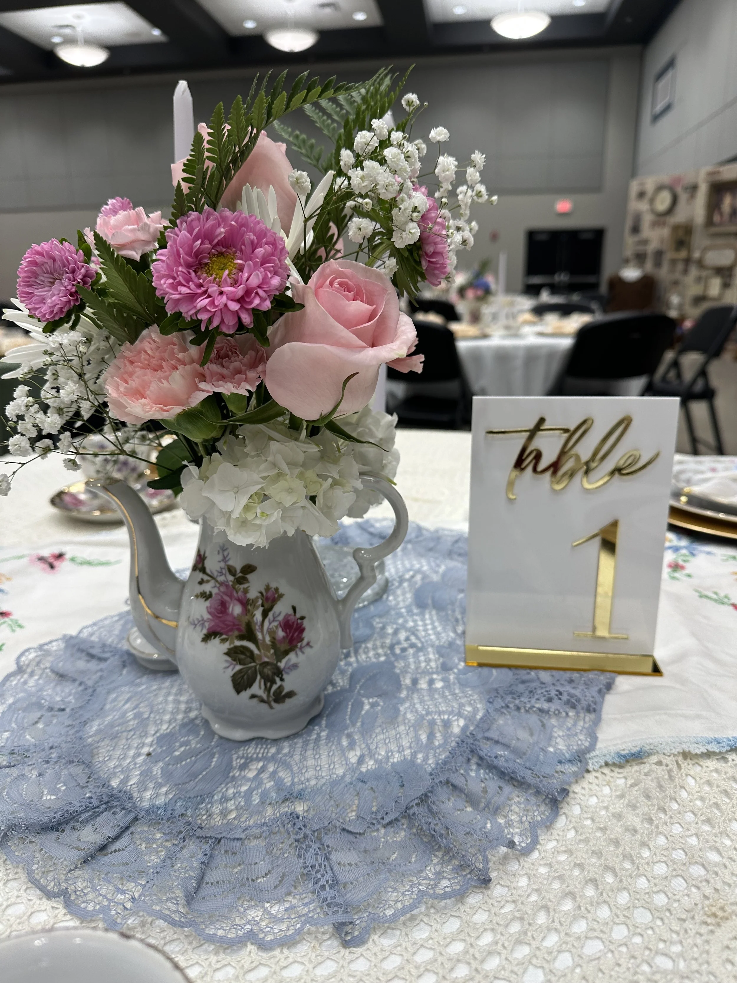 A floral centerpiece with pink, white, and purple flowers in a decorative teapot vase sitting on a table with lace tablecloth. A table number sign with the number 1 is also on the table, indicating a table setting at a formal event.