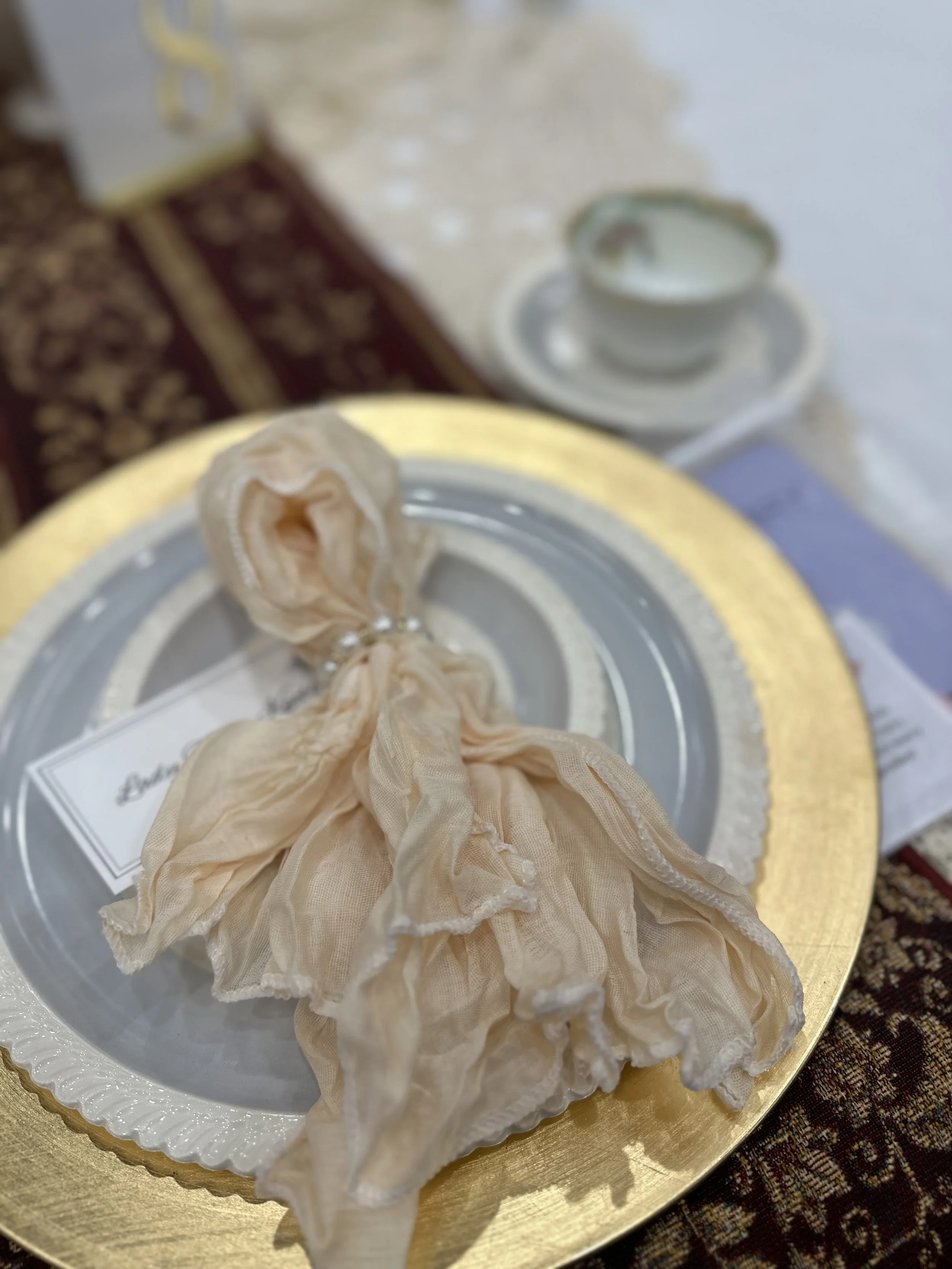 A decorative gift box with a beige fabric bow on top, placed on a gold-rimmed plate at a table. In the background, there is a white teacup on a matching saucer and a small metal sign with the word 'Sweet' in gold letters.