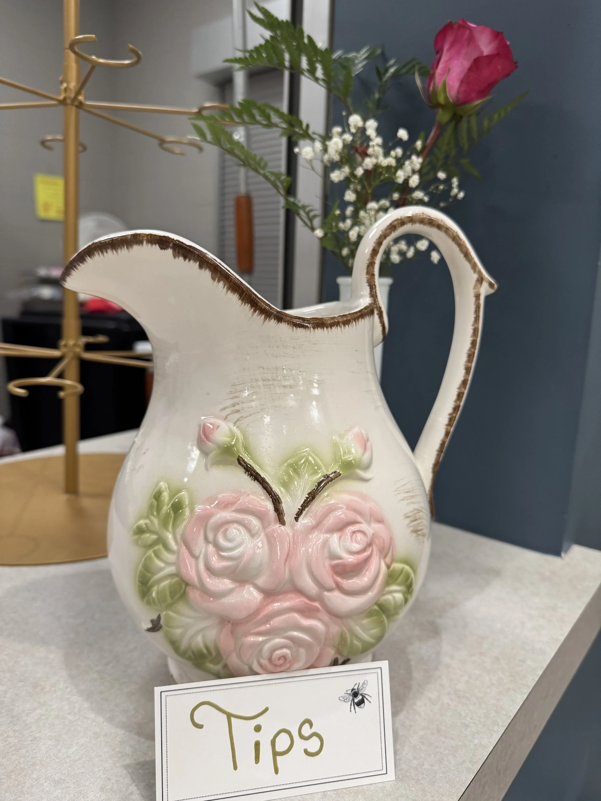 A ceramic teapot with pink roses and green leaves design, placed on a countertop. There is a small sign with the word 'Tips' and a bee illustration in front of it. In the background, a vase with a pink rose, white baby's breath, and greenery is visib