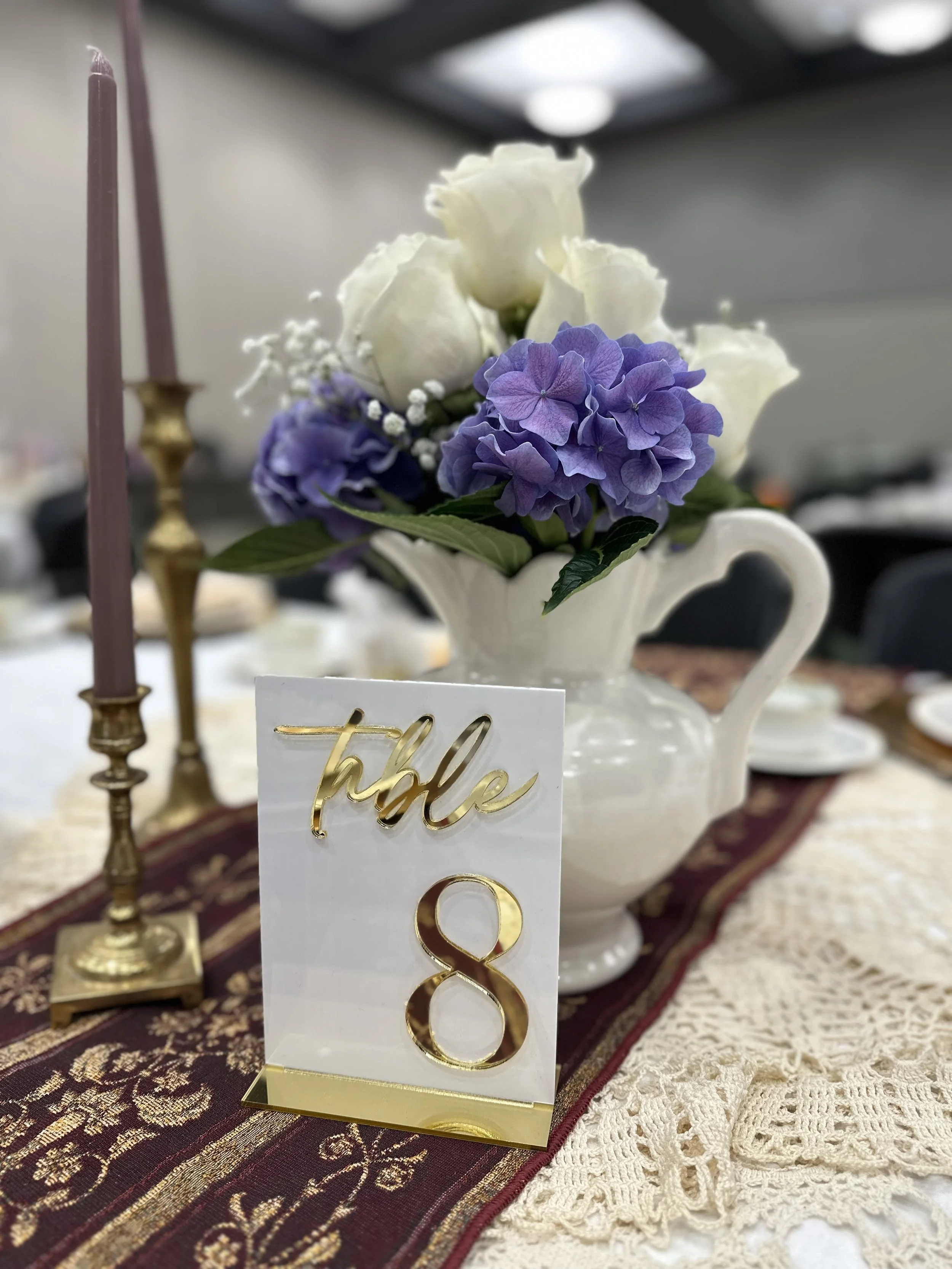 A table centerpiece with white and purple flowers in a white ceramic pitcher, gold candlesticks with pink candles, and a table number sign reading 'table 8' in gold lettering, on a lace and burgundy table runner.