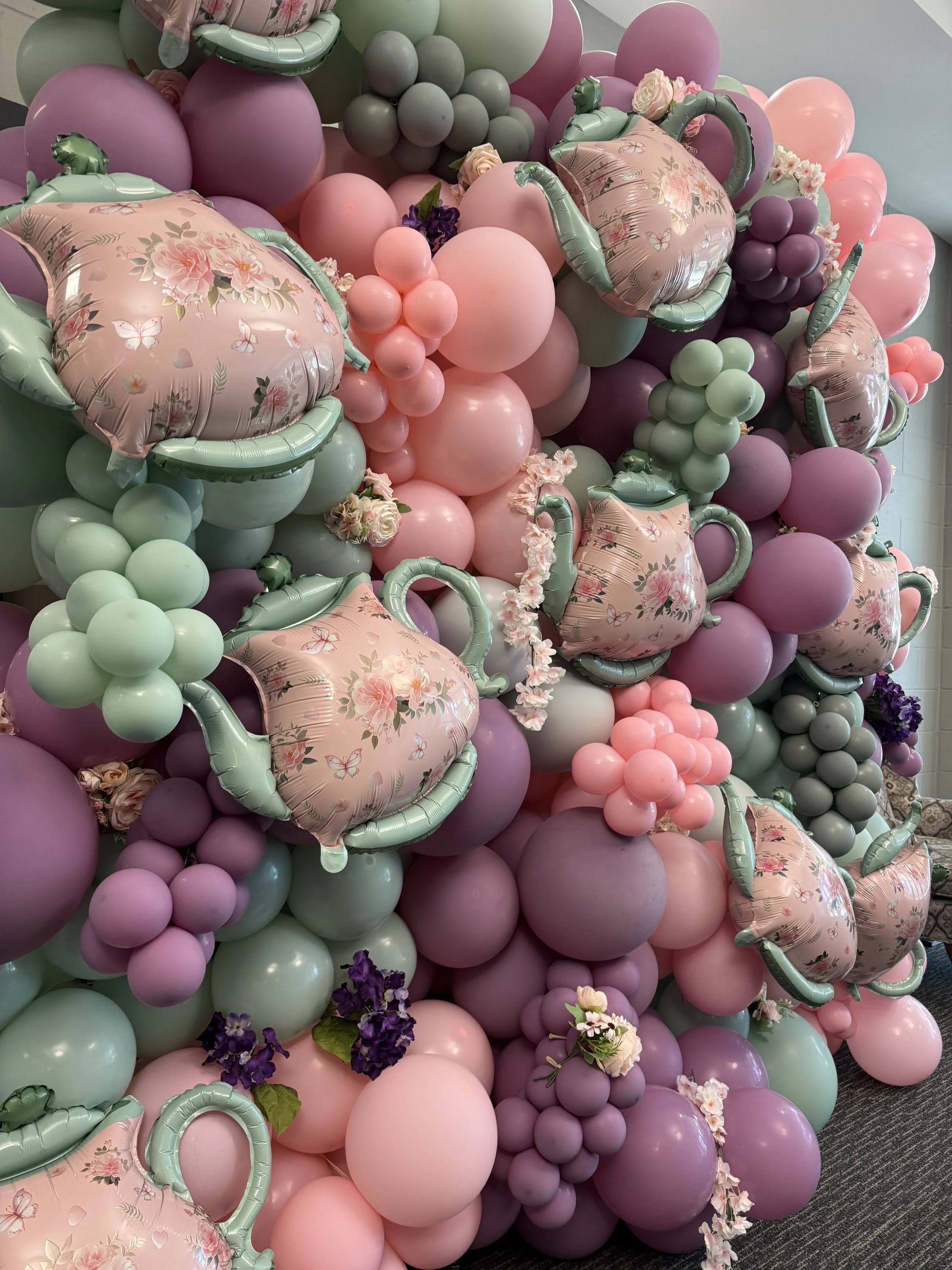 A colorful balloon decoration with pink, purple, mint green, and gray balloons, featuring teapot-shaped balloons, flower accents, and grape-like clusters.
