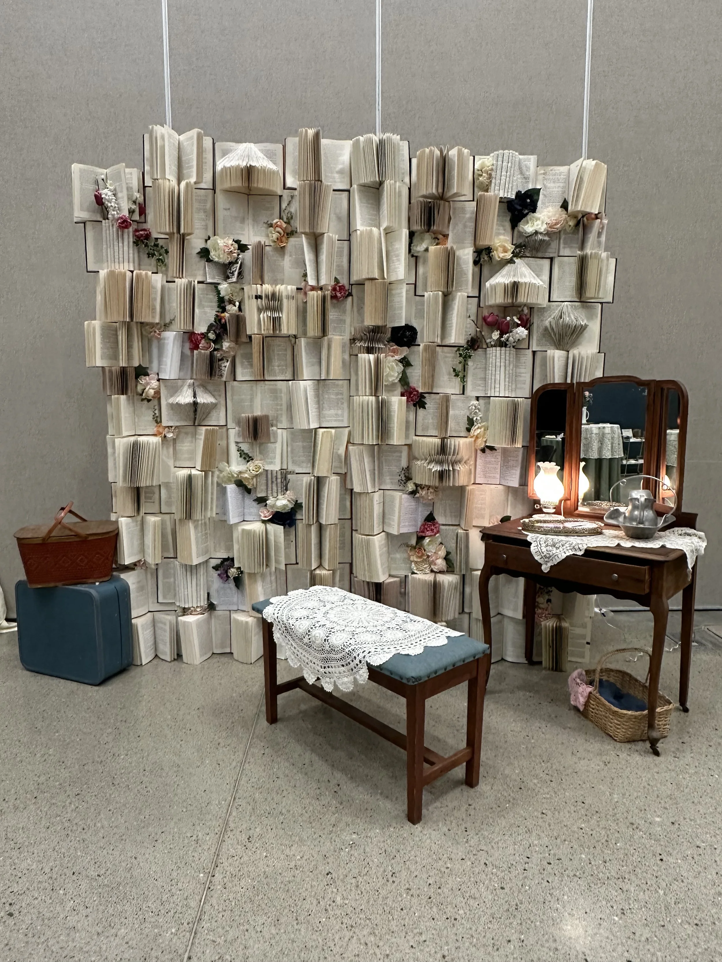 A decorative wall made of open books arranged vertically and horizontally, adorned with artificial flowers. In front, a vintage wooden vanity with a three-panel mirror, lace doily, lamp, and teapot. A wooden stool with a crochet cover is nearby, alon