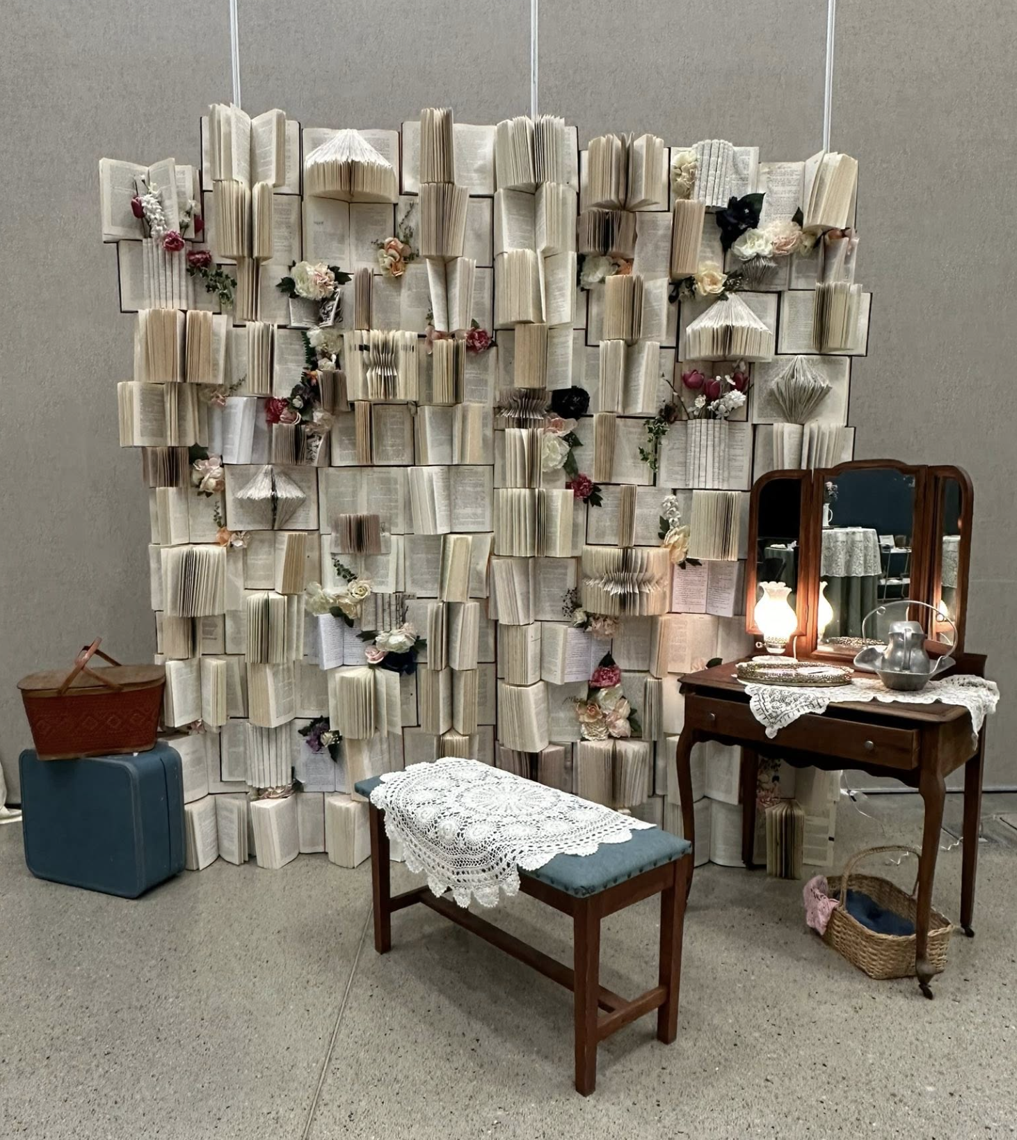 A decorative display of open books arranged on a wall with flowers, a vintage vanity with a mirror, a lace-covered bench, and various vintage accessories.