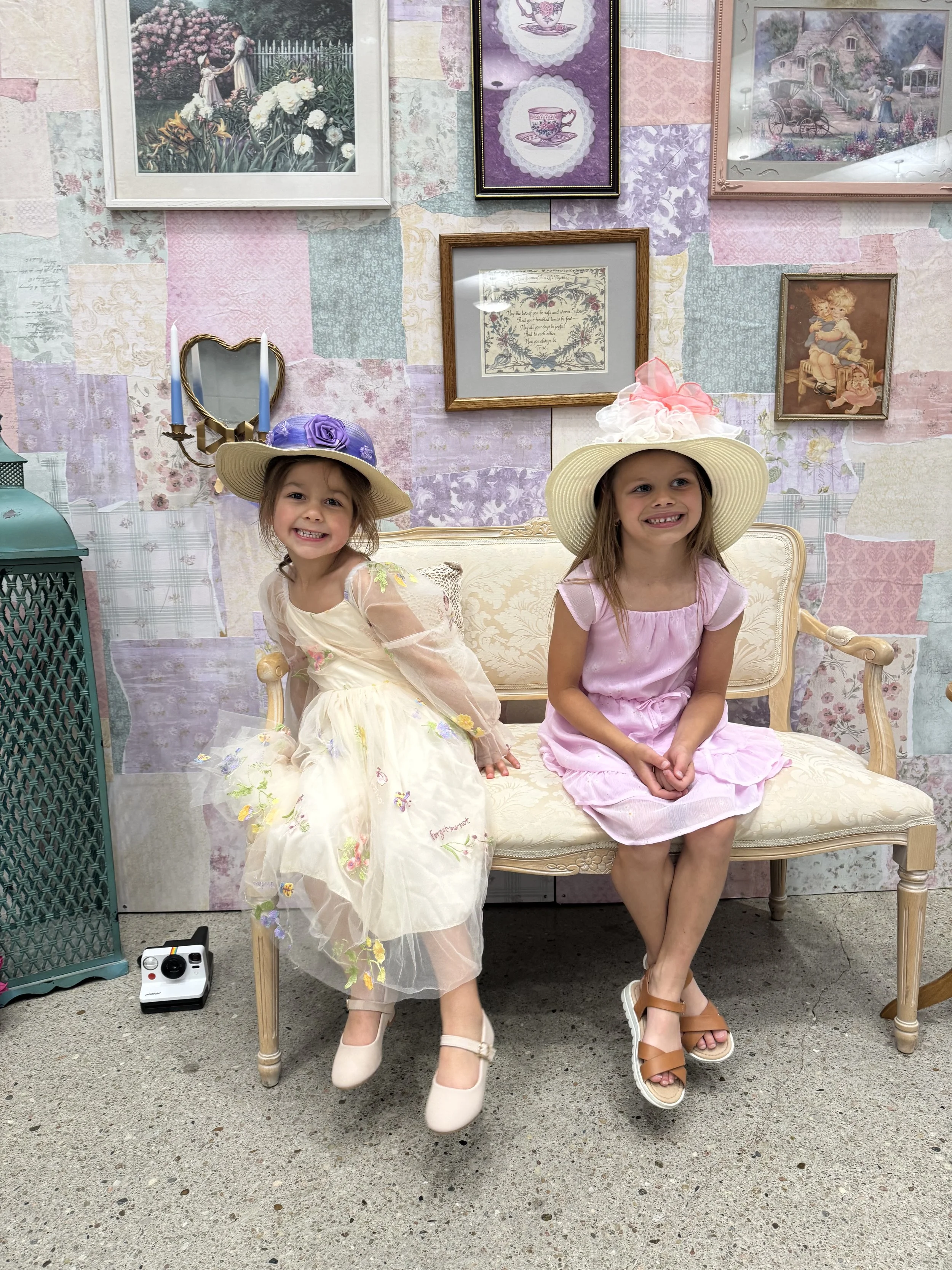 Two young girls wearing pastel-colored dresses and large wide-brimmed hats sitting on a vintage cream-colored sofa. They are smiling and posing in front of a colorful patchwork wall decorated with framed pictures and artwork.