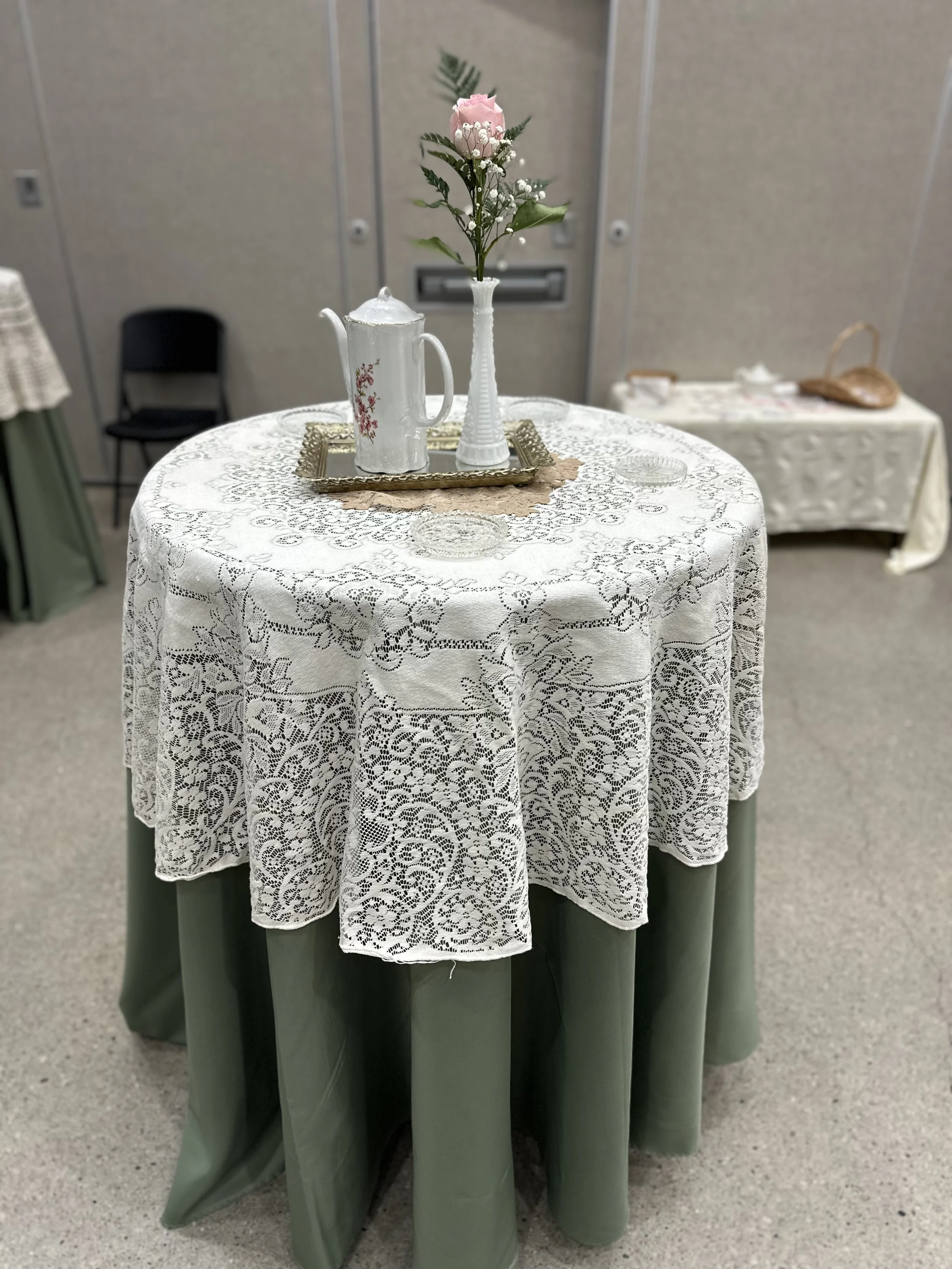 A round table with a green tablecloth covered by a lace overlay, decorated with a vase of pink roses and white baby's breath, a silver tray, and a white teapot. In the background, there are other tables and chairs.
