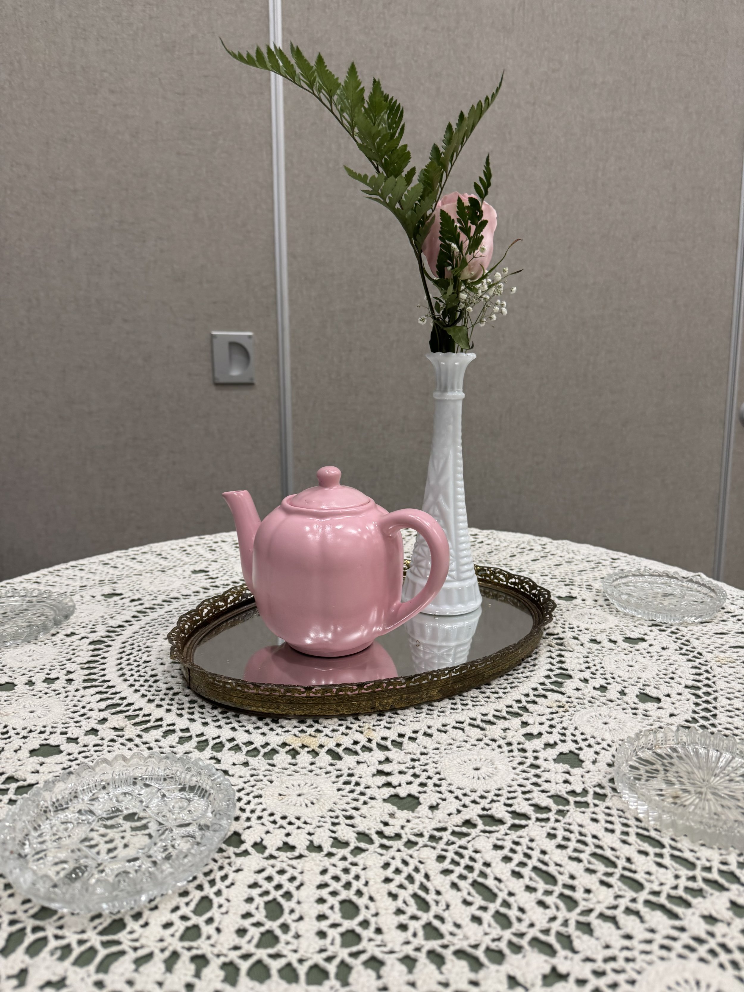 A table with a lace tablecloth, featuring a pink teapot on a silver tray, a tall white vase with green leaves and pink flower, and five glass bowls around the edges.