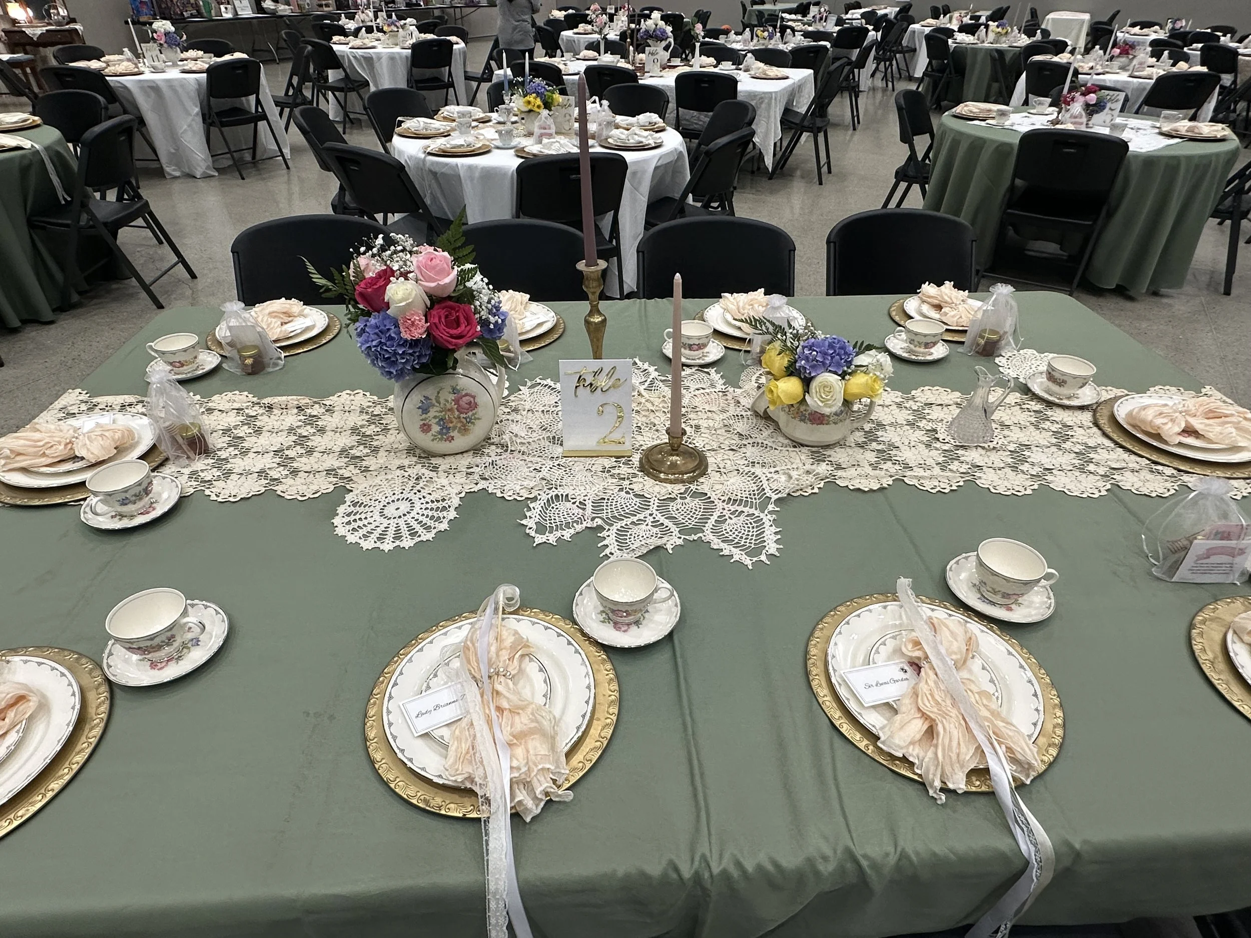 Round table decorated with a lace table runner, floral centerpieces with roses, hydrangeas, and other flowers, gold-rimmed plates, teacups, and napkins, set for a formal event or wedding reception.