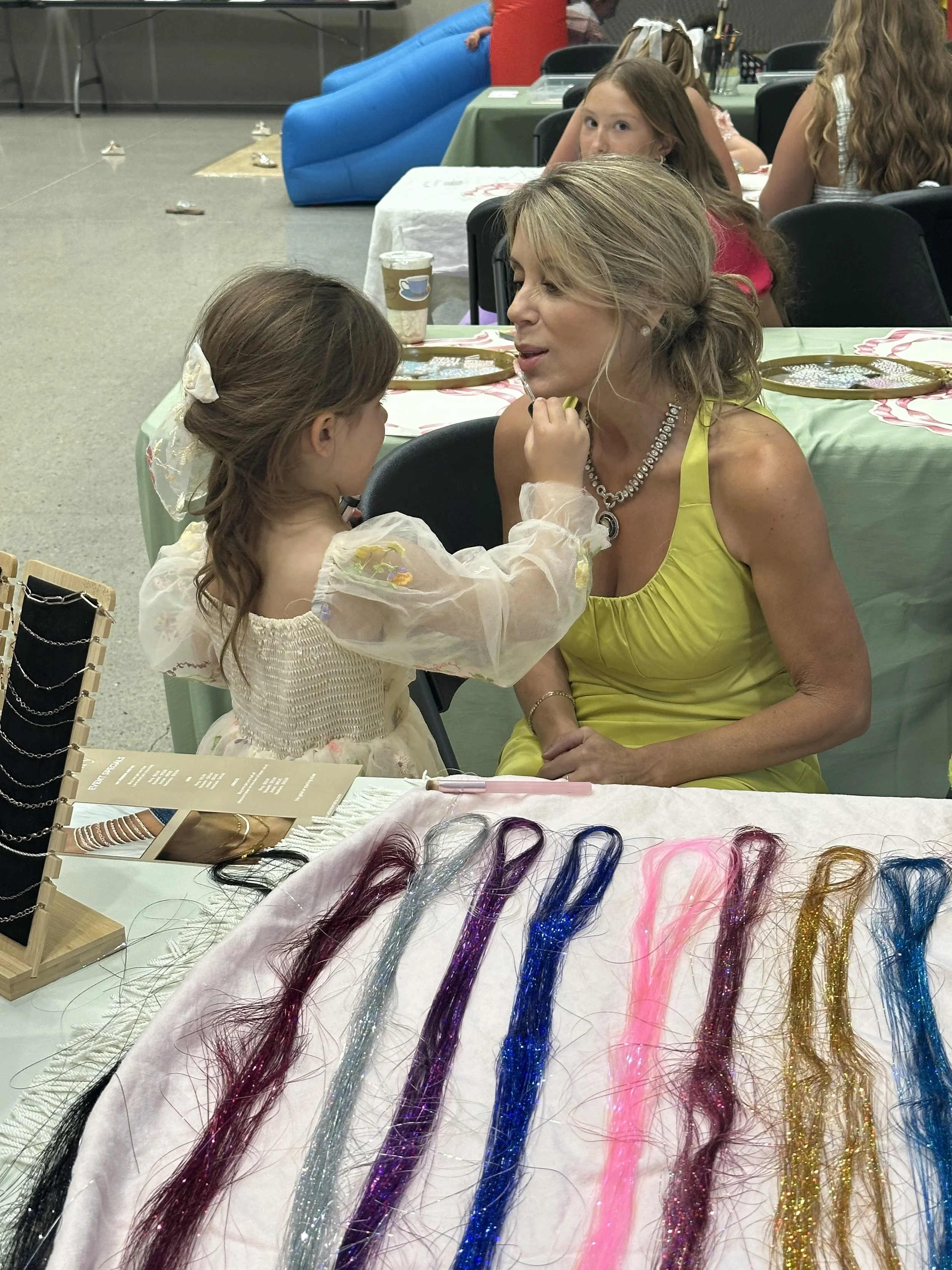 A young girl with long hair and a bow in her hair is touching a woman's face at a table during a craft or jewelry event. The woman is wearing a yellow dress and a necklace. There are colorful hair extensions laid out on the table in front of them, wi