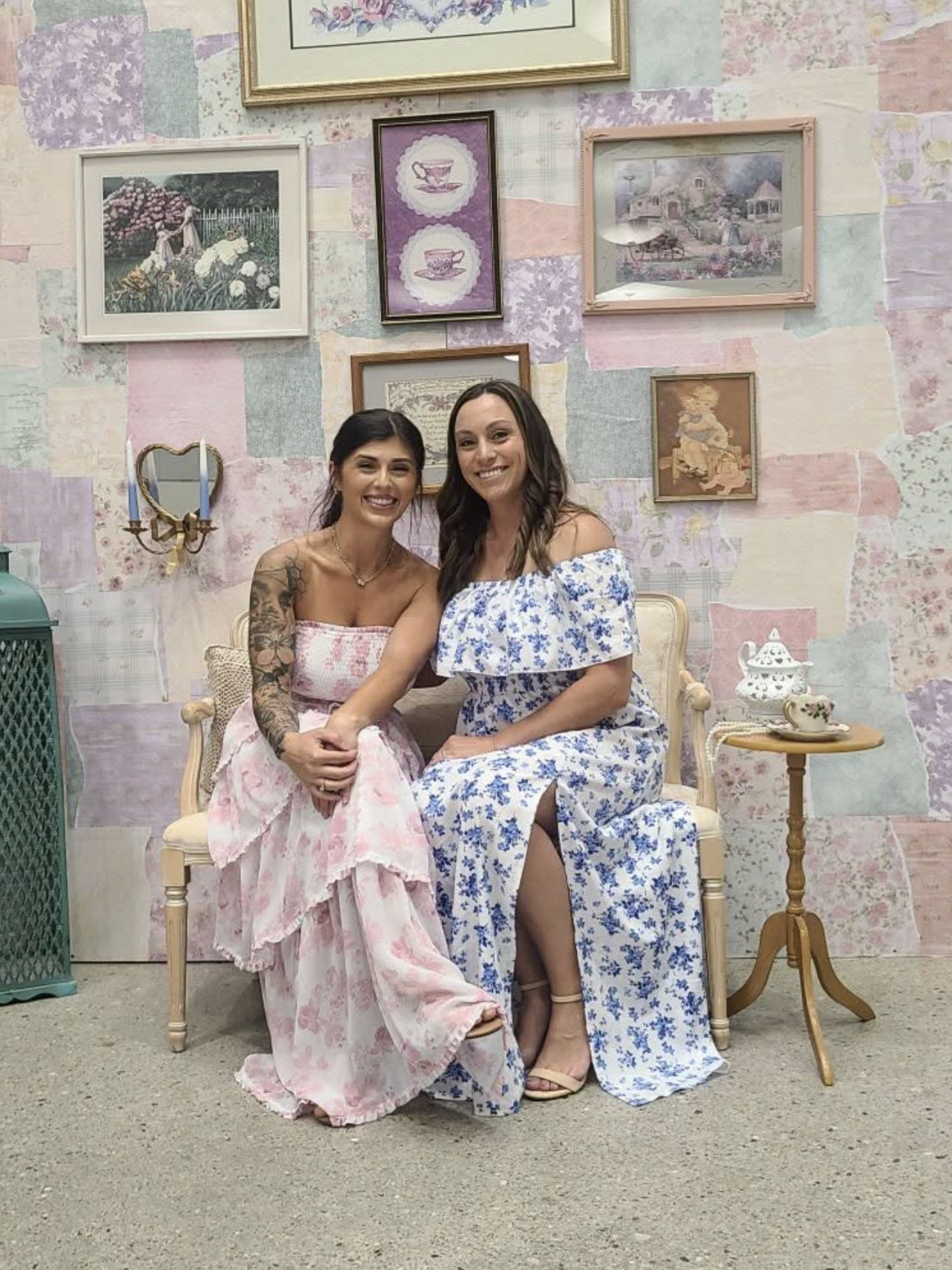 Two women sitting on a vintage-style bench, smiling, with a pastel-colored wall decorated with framed artwork behind them. One woman wears a pink floral dress with tattoos on her arm, and the other wears a blue floral off-shoulder dress.