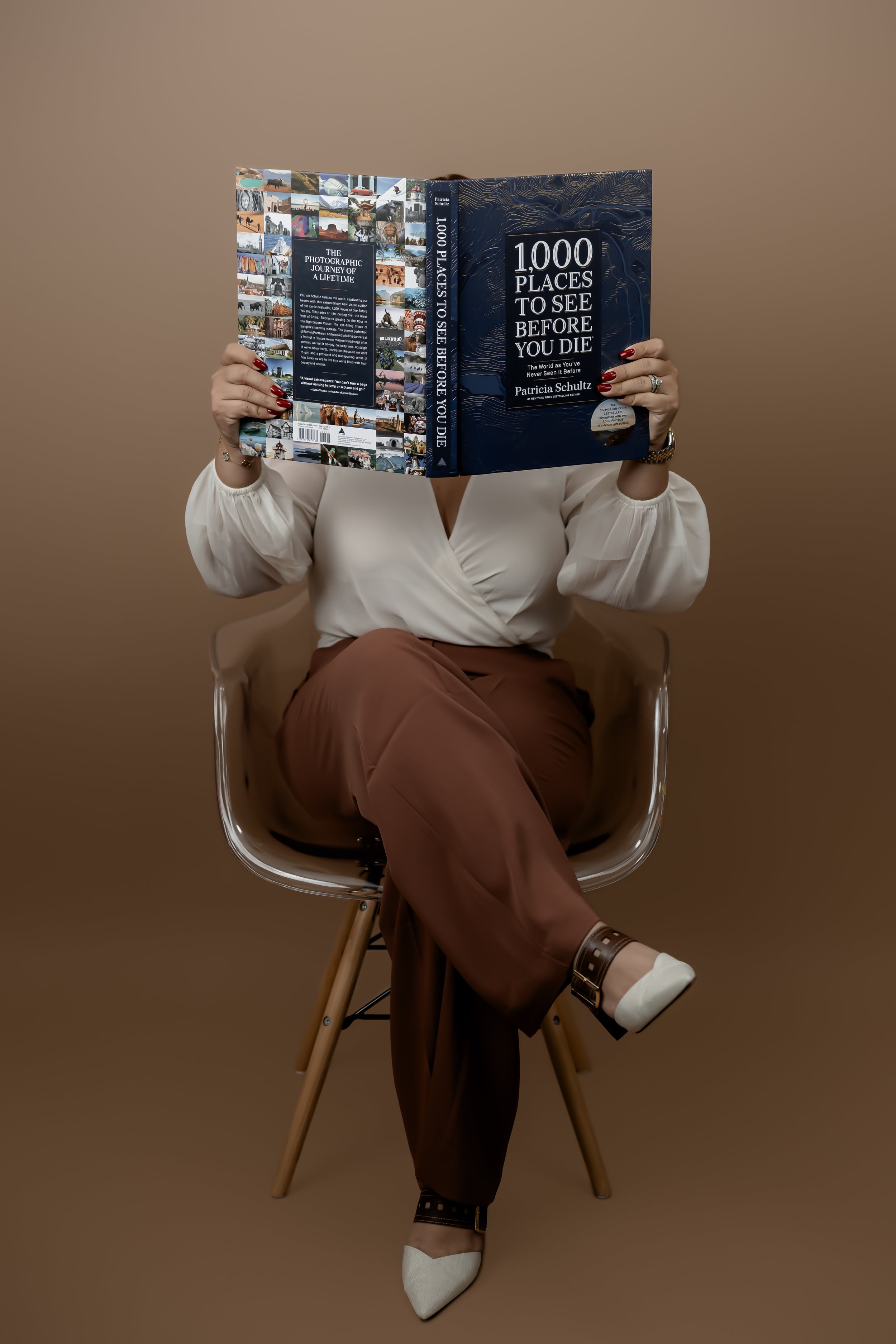 A woman sitting on a modern chair with wooden legs, reading a large book titled '1000 Places to See Before You Die.' She is wearing a white blouse, brown pants, and white shoes with a buckle. Her face is hidden by the book, and her fingernails are painted red, with a ring on her right hand.