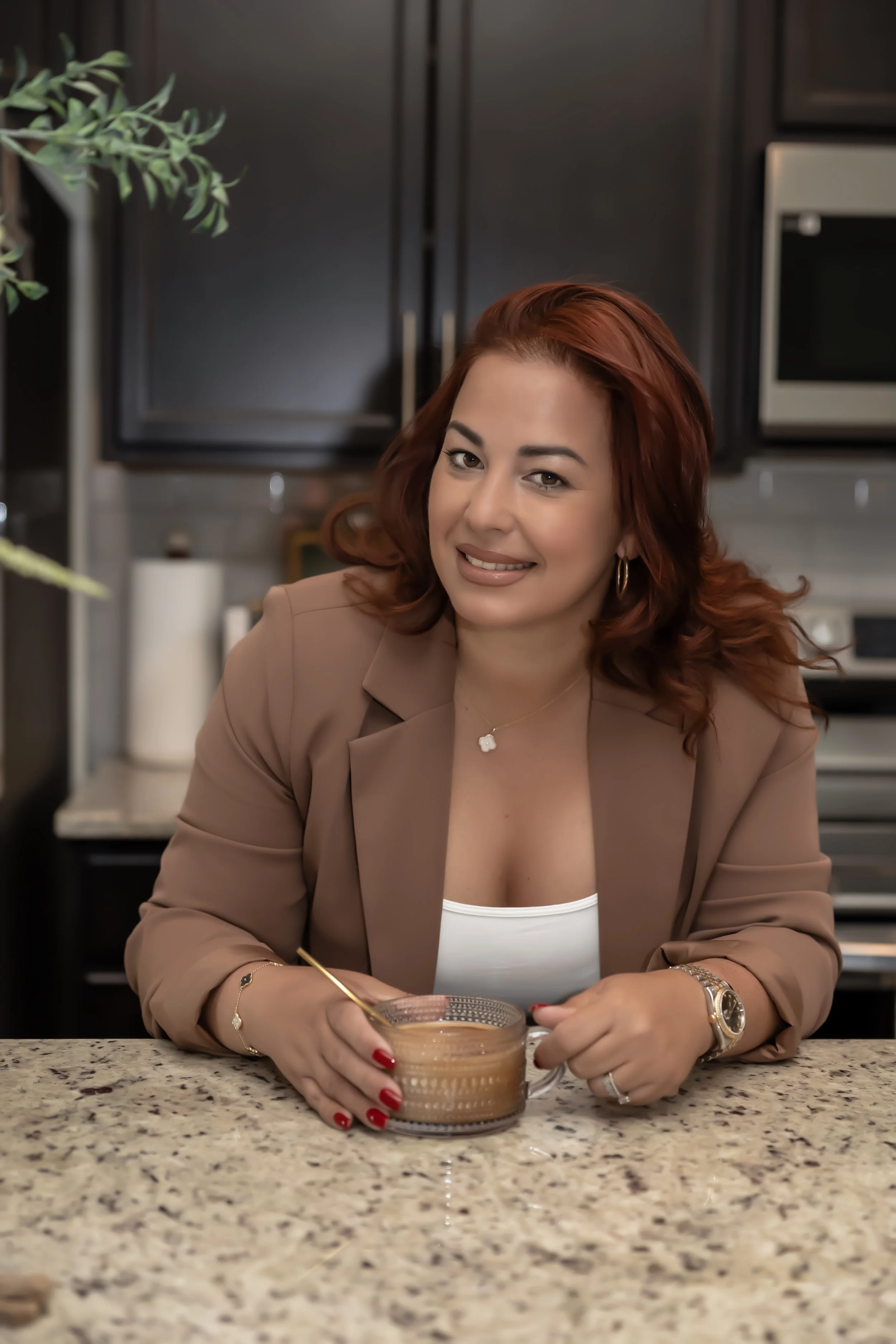 A woman with red hair, wearing a beige blazer and white top, sitting at a kitchen counter with a cup of coffee, smiling at the camera.