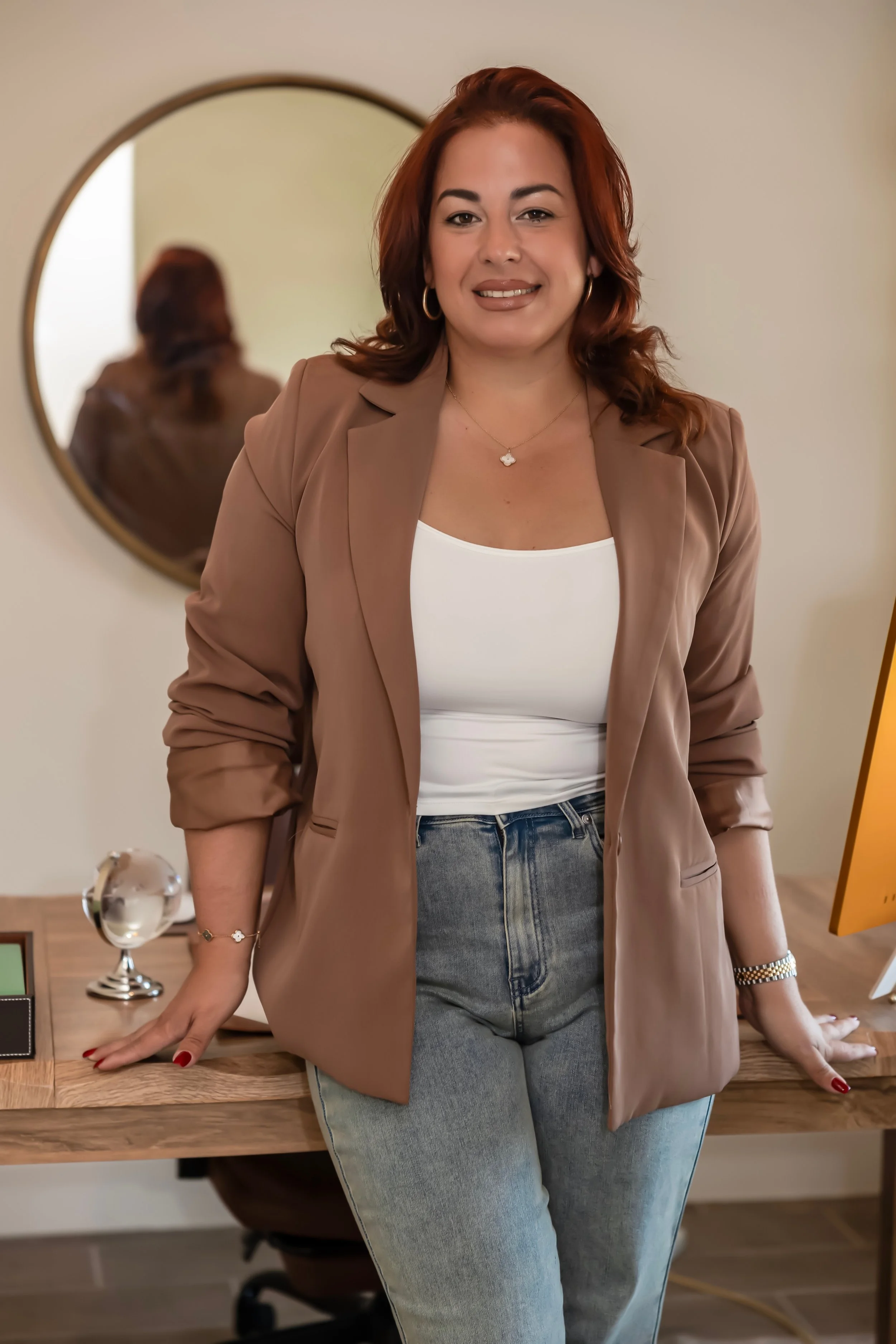 A woman with red hair, wearing a tan blazer, white top, and jeans, standing in front of a desk with a mirror behind her reflecting her back.