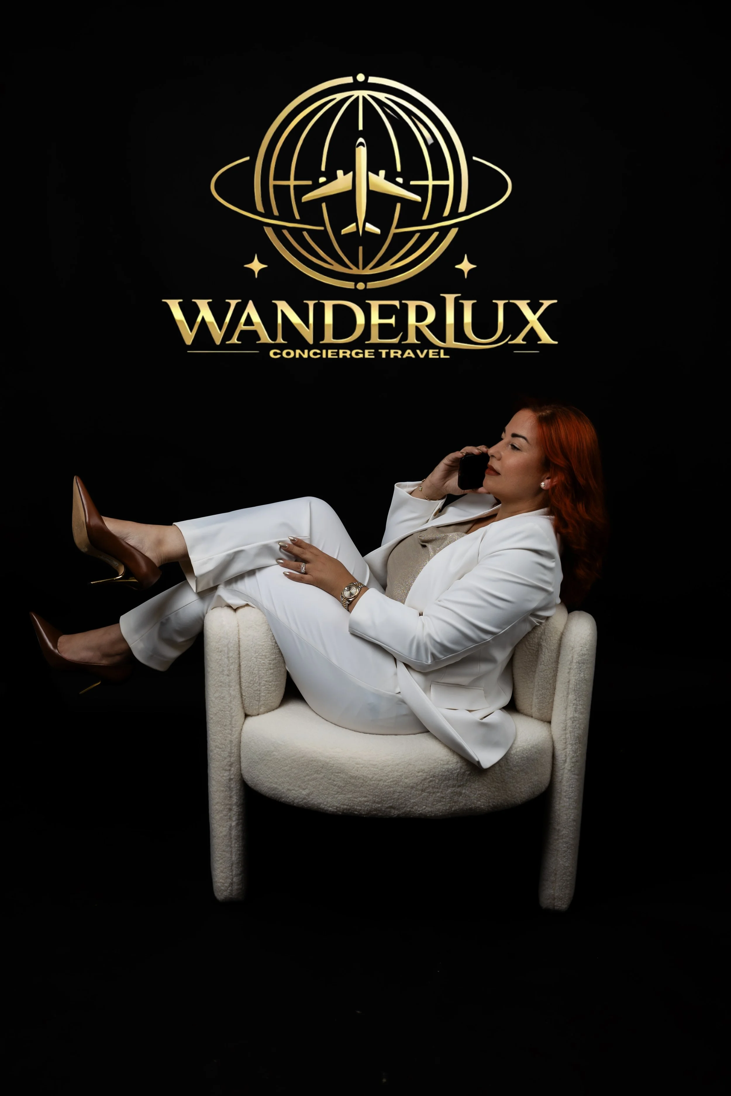 A woman with red hair, wearing a white suit and brown high-heeled shoes, reclining in a cream-colored armchair while talking on a cellphone, with a black background and a gold logo that reads "WanderLux Concierge Travel" above her.