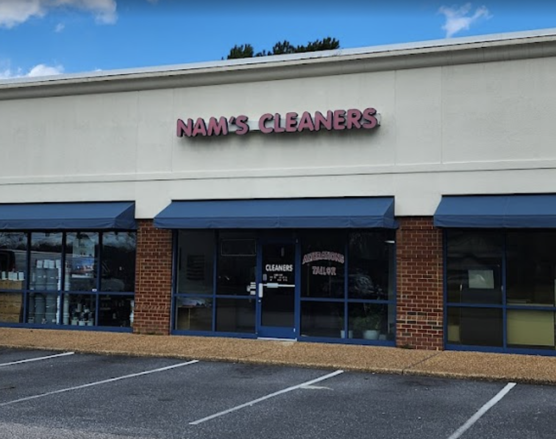 Front of a dry cleaning store called 'Nam's Cleaners' with a sign above the entrance, blue awnings, and empty parking spaces in front.