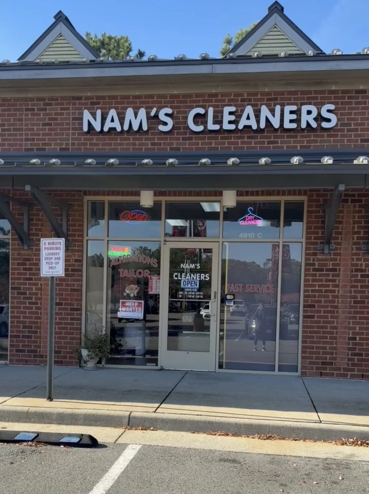 Front of a brick building with a sign that reads 'NAM'S CLEANERS'. The glass door has signs including 'Open' and 'Dry Cleaners'. Inside, there are more signs, and the reflection of the parking lot and people outside can be seen in the windows.