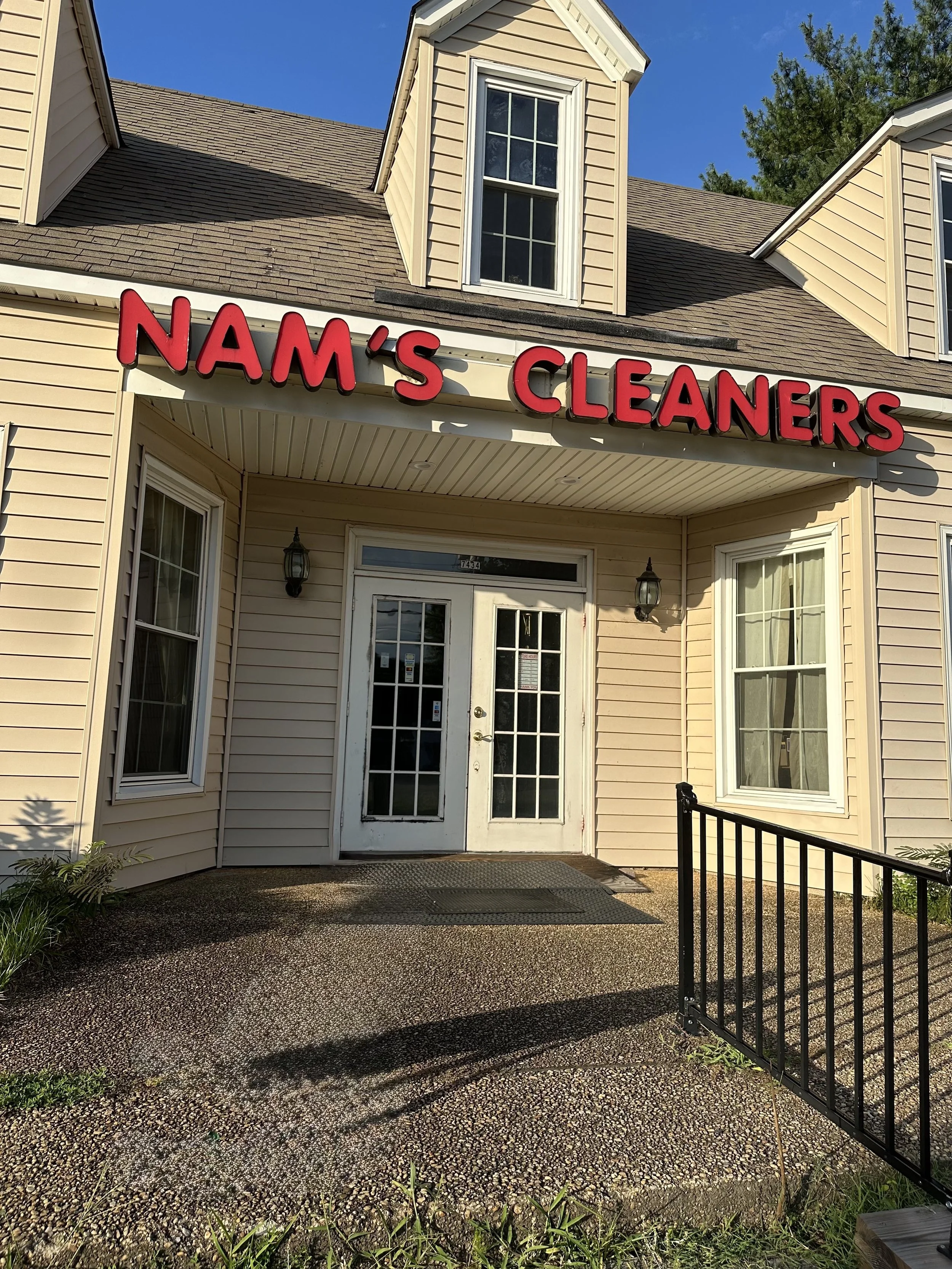 Front entrance of a beige building with a sign reading 'NAM'S CLEANERS' in red letters. French doors, two windows, and two wall-mounted lights are visible.