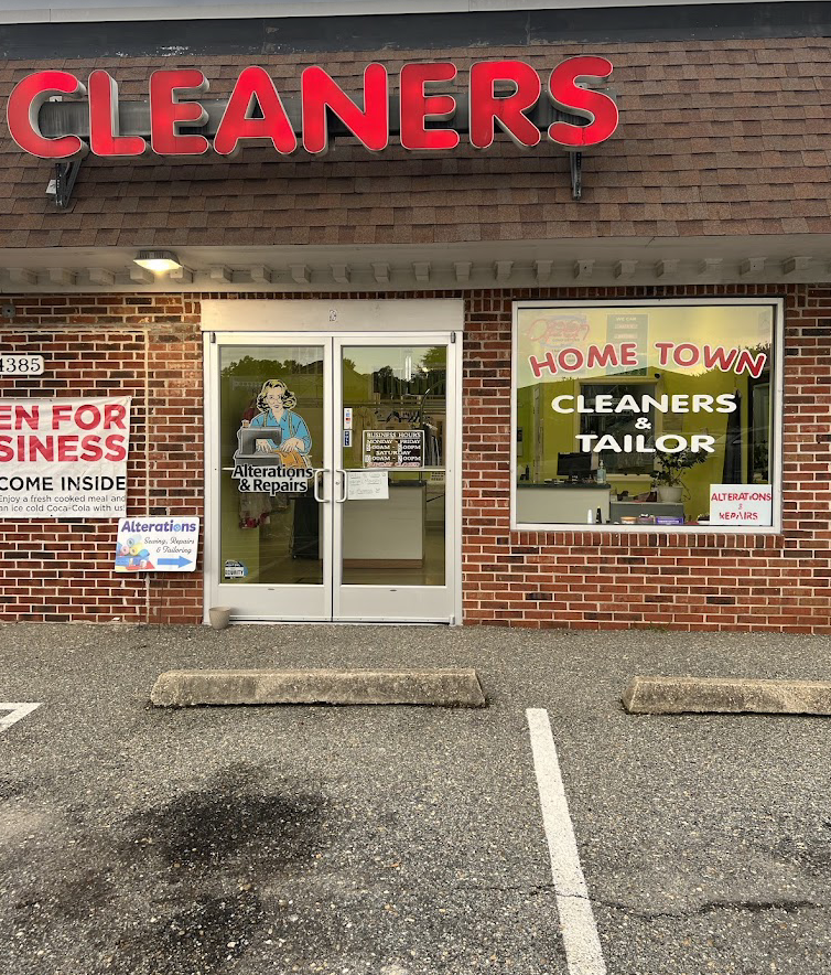 Exterior view of a dry cleaner shop called Home Town Cleaners & Tailor, with red sign reading 'CLEANERS' above the entrance, a glass door with a woman illustration and business hours, and posters advertising alterations and repairs. There is a parking lot with two empty spaces in front.