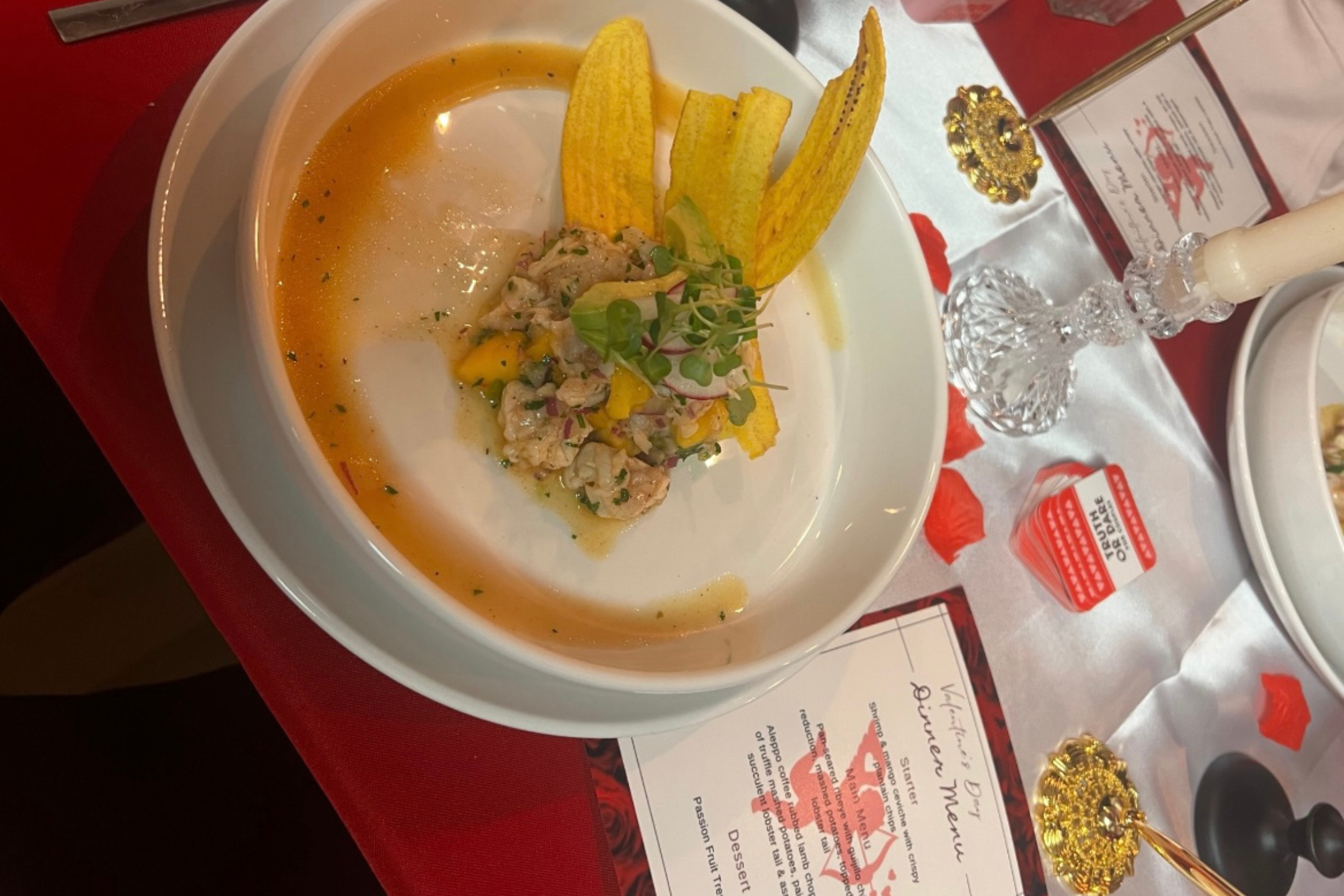 Bowl of spicy seafood soup garnished with microgreens and plantains, set on a red table with a decorative candle and menu.