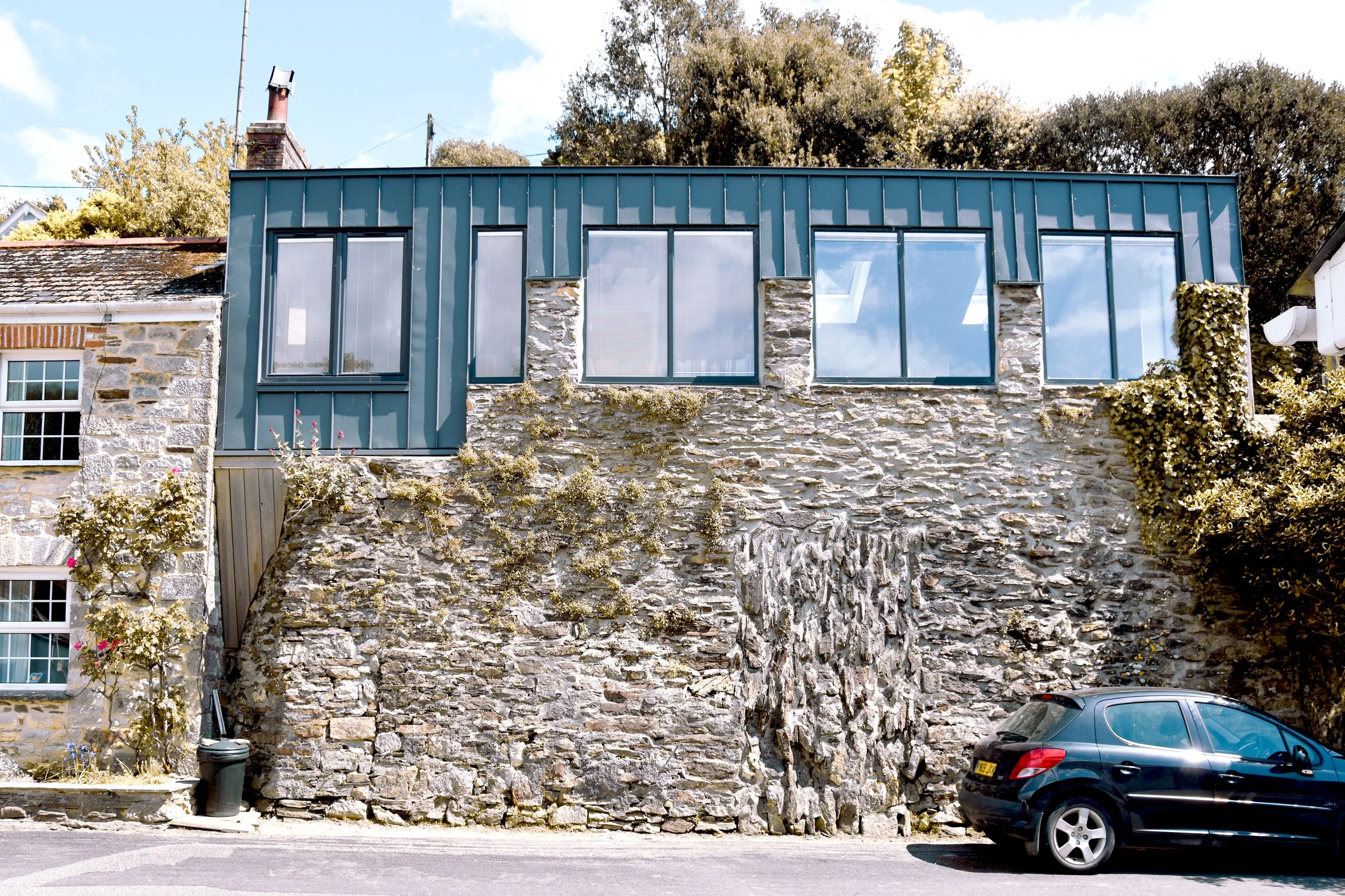 A house with a stone foundation and modern blue metal siding on the upper level, featuring large windows, with a black car parked in front on a sunny day.