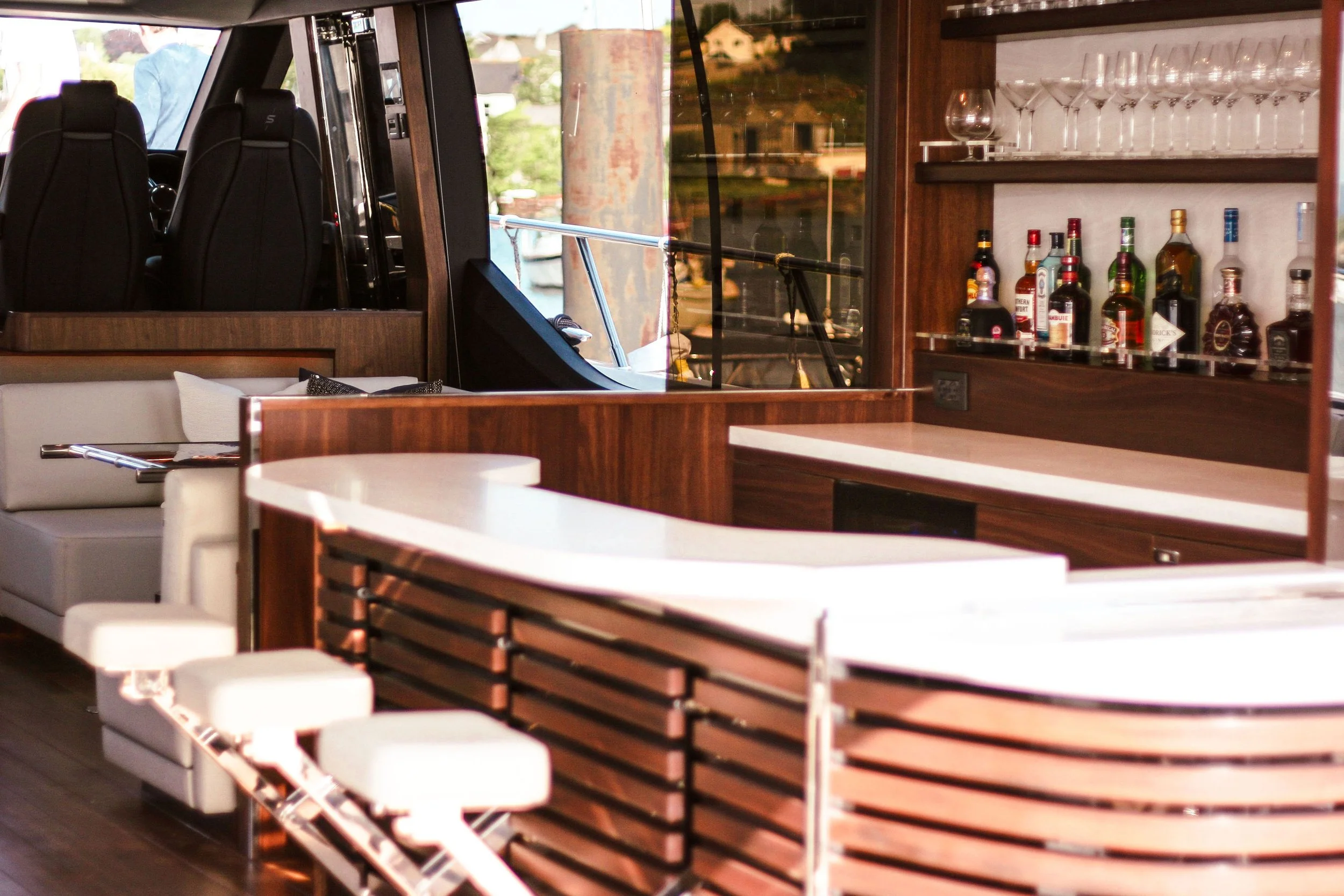 Interior of a boat bar with a white L-shaped sofa, stools, and a wooden bar counter with liquor bottles and glasses, with boat seats visible through a window.