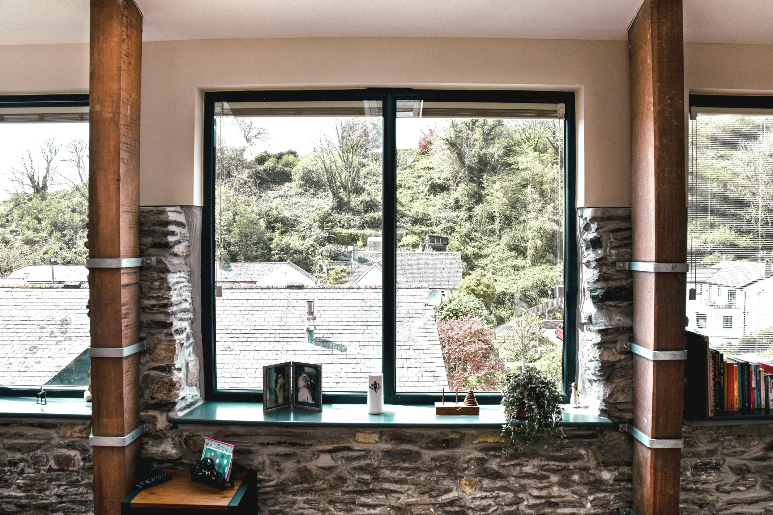 View through a large window showing rooftops and trees, with objects like framed photos, a candle, and a plant on the windowsill inside a room with stone walls and wooden beams.