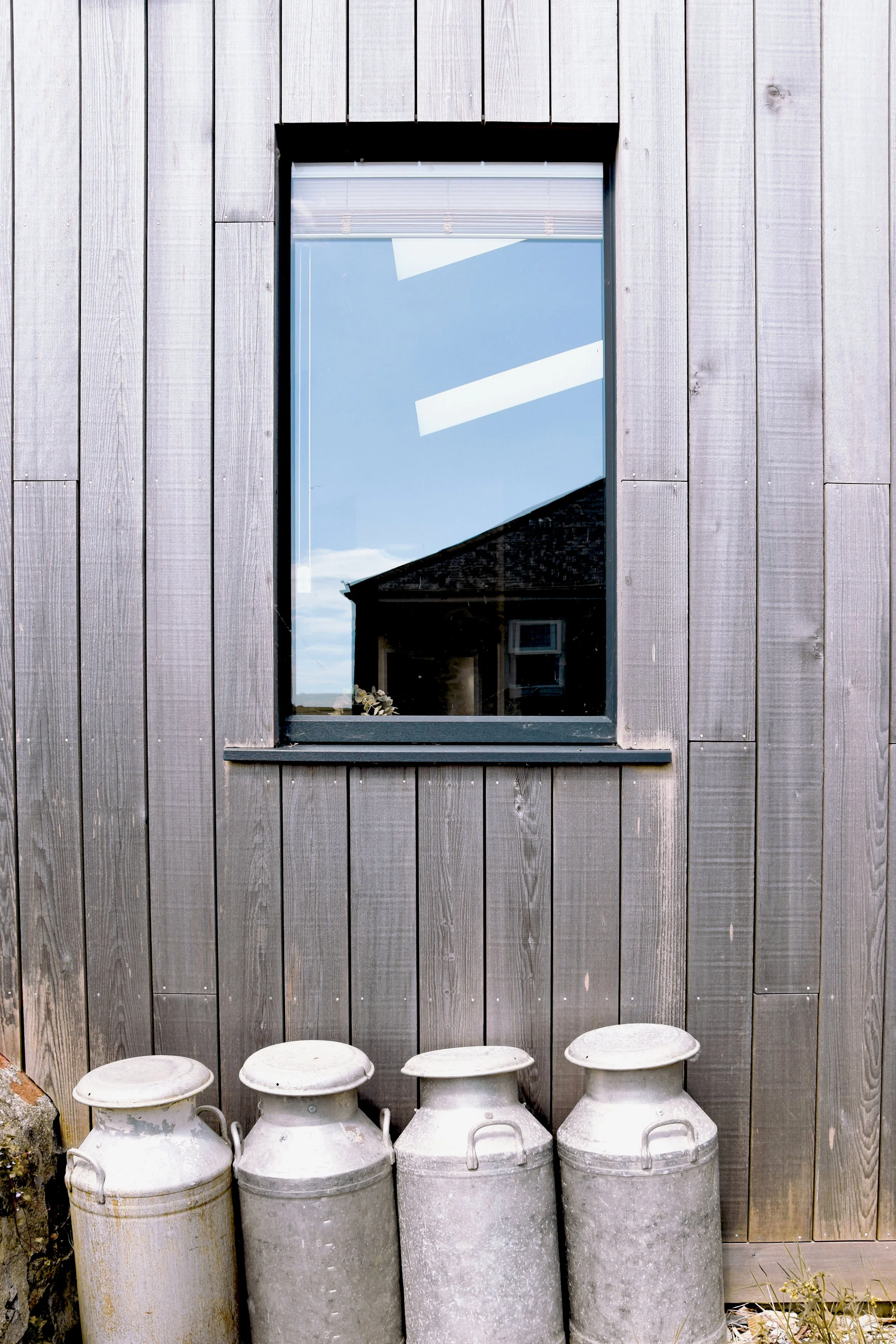Four antique milk cans placed outside a building with a window on a wooden wall.
