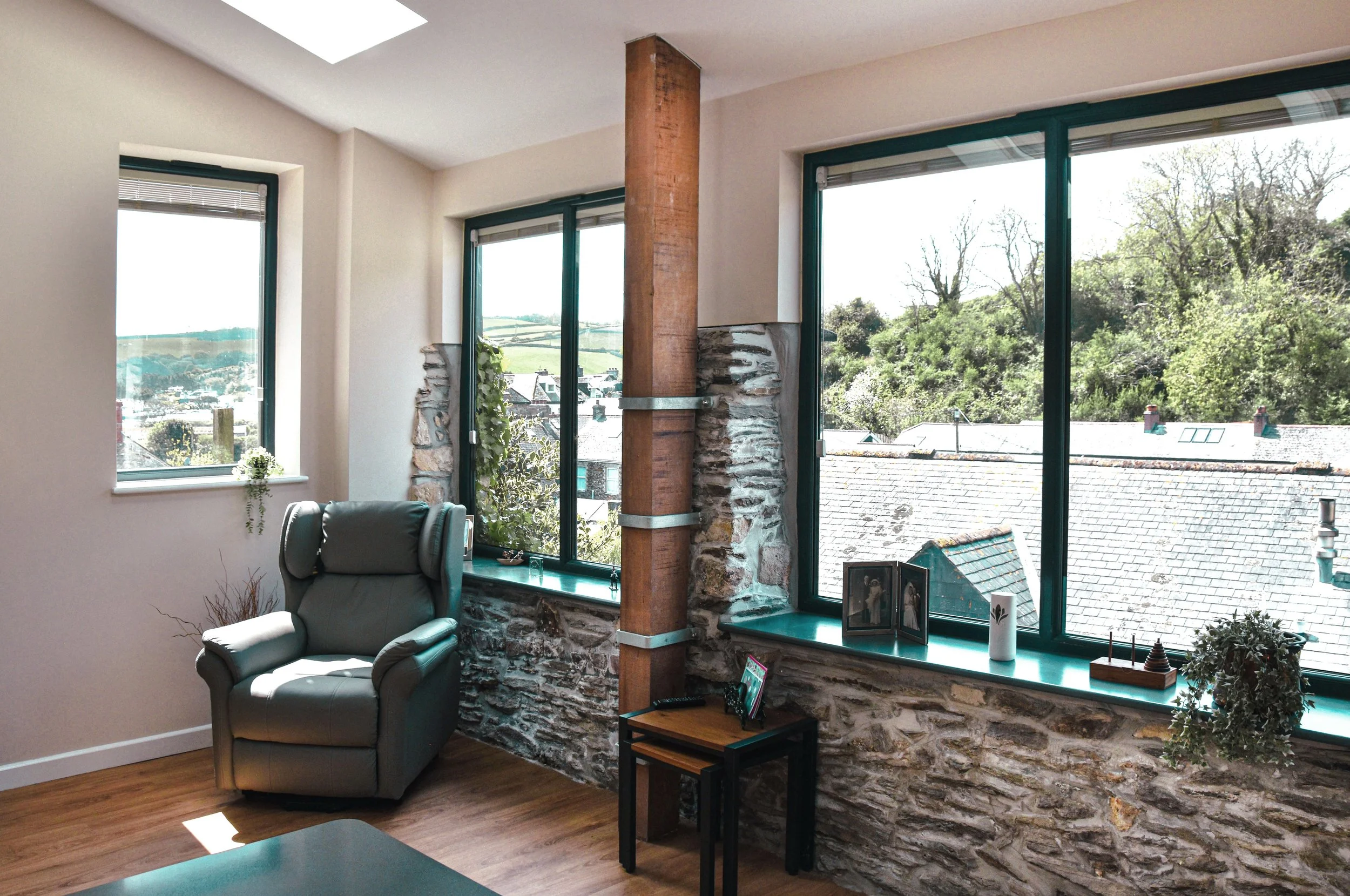 Interior view of a room with large windows overlooking a hilly landscape, decorated with a black leather recliner, small black side table, and various decorative items on the windowsill.