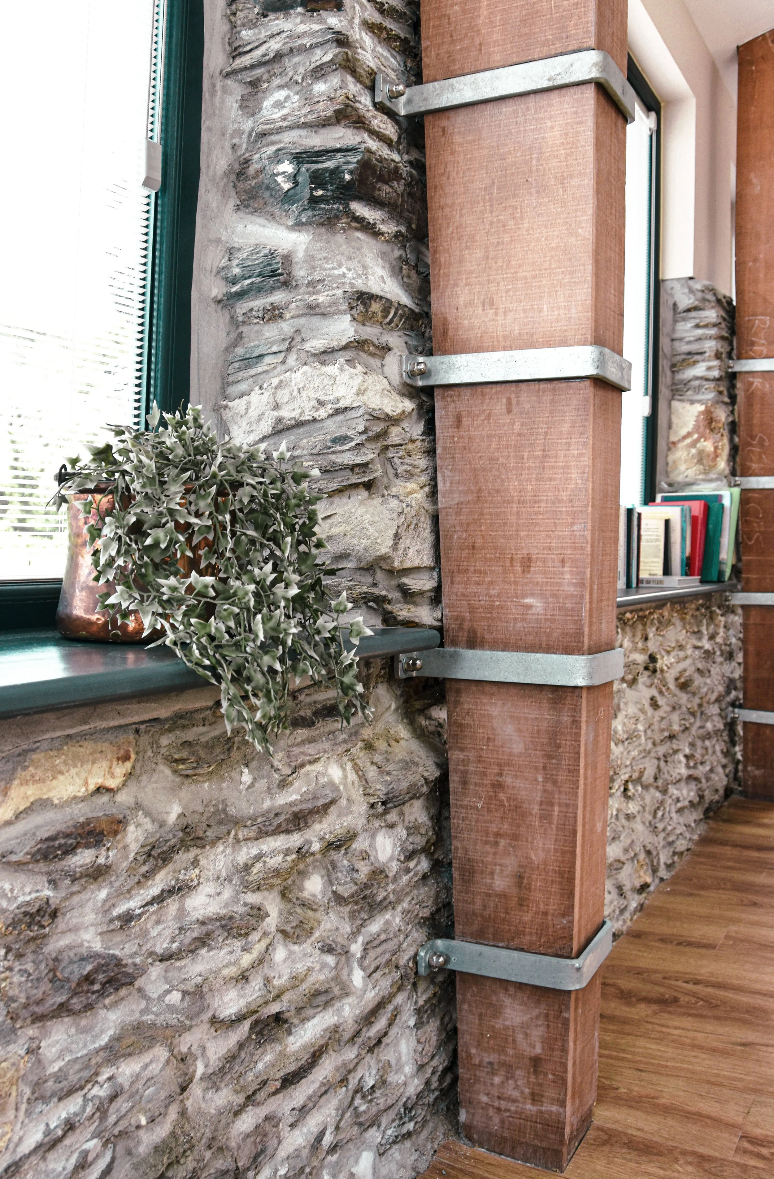 Close-up of an interior wall with a large wooden support beam held by metal brackets, a stone wall, and a window sill with a potted plant and books.