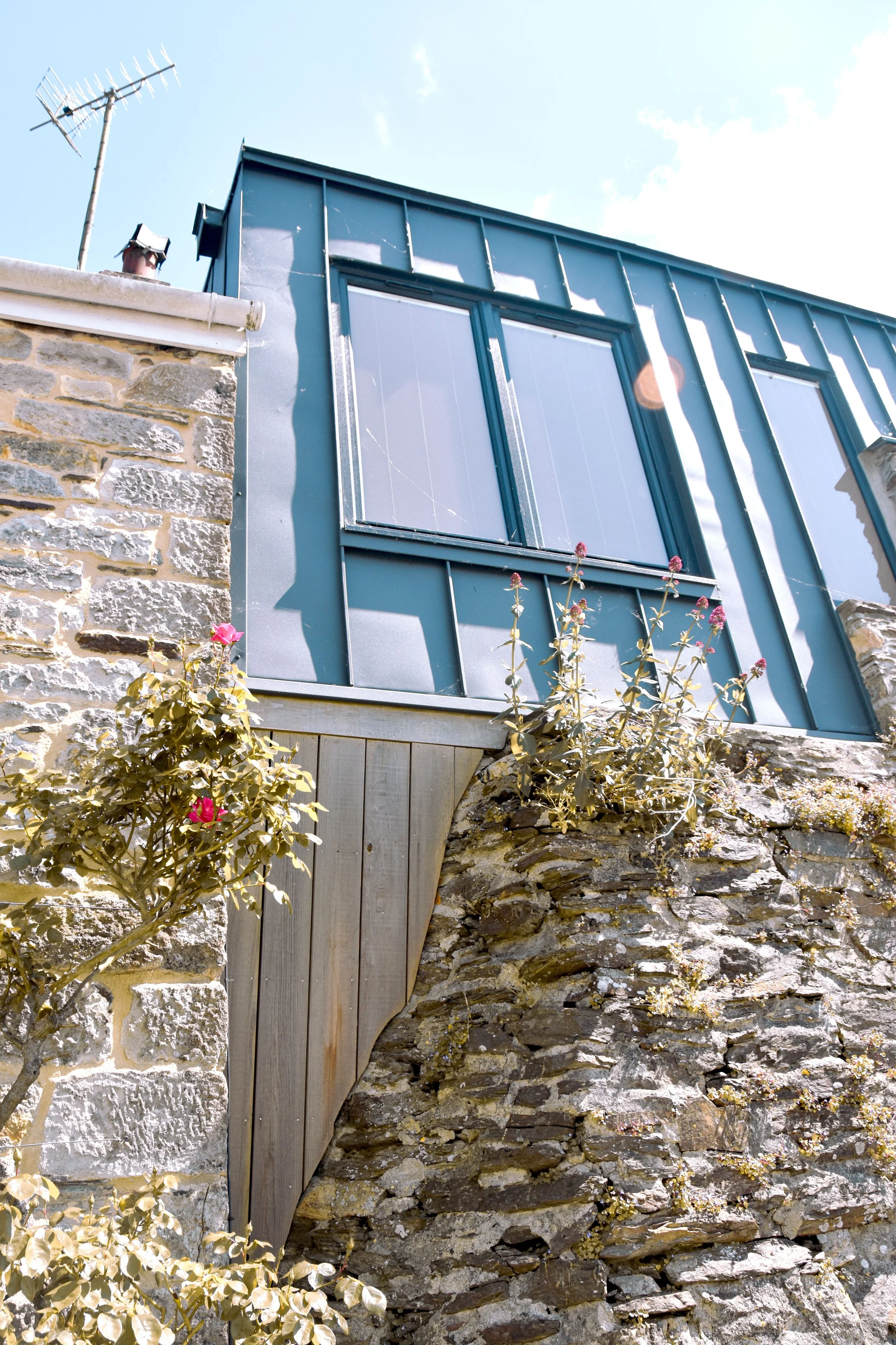 A house with stone and blue metal siding exterior, large window with closed blinds, and flowering plants growing along the stone wall, under a clear blue sky.