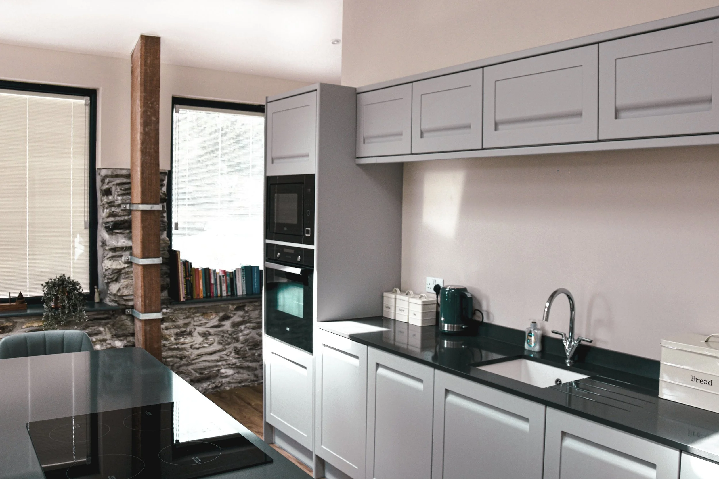 Modern kitchen with white cabinets, black countertop, built-in oven and microwave, sink, and appliances. There are large windows with blinds and a wood and stone accent wall.