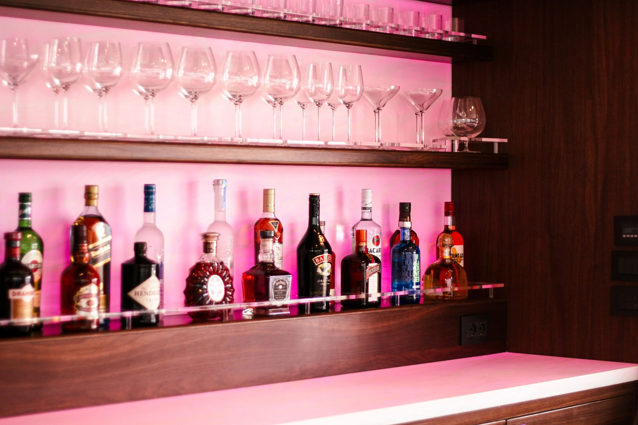 A bar shelf with pink backlighting displaying various liquor bottles and empty glassware.