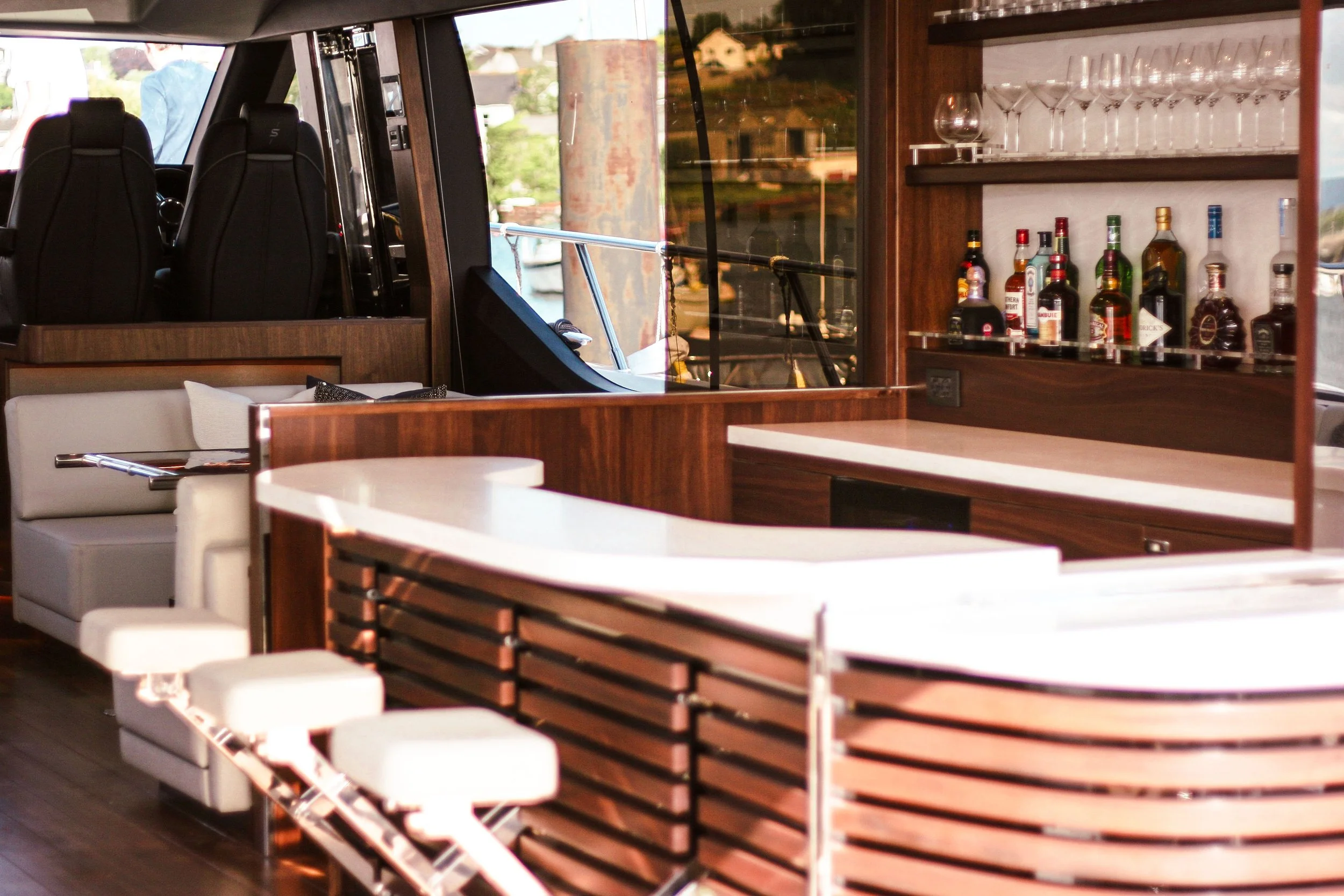 Interior of a boat bar featuring a curved white counter, a shelf with various alcohol bottles, glassware, and plush seating with white cushions.