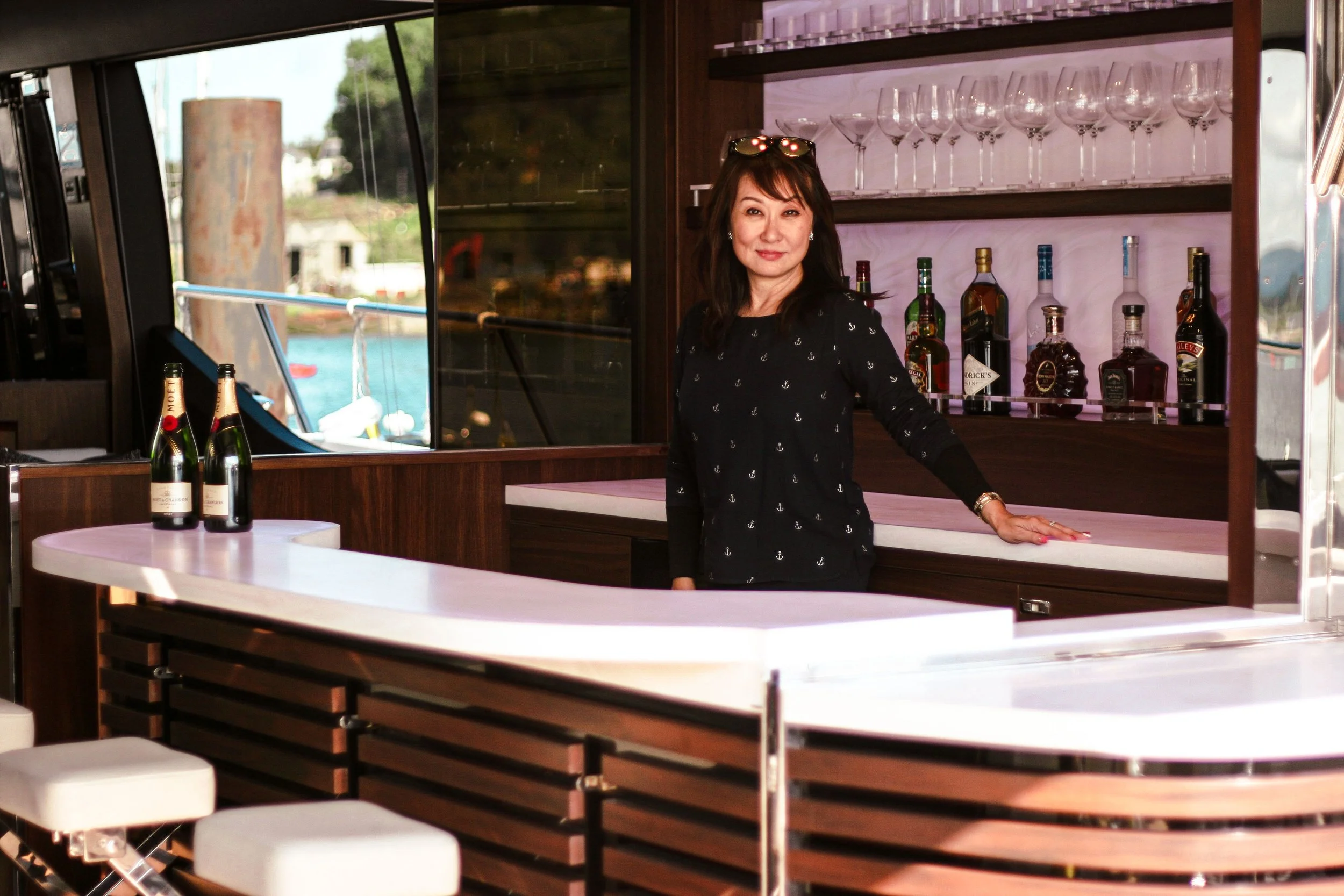 A woman standing behind a bar on a boat, with bottles of alcohol and wine glasses behind her, and two bottles of sparkling wine on a white curved countertop.