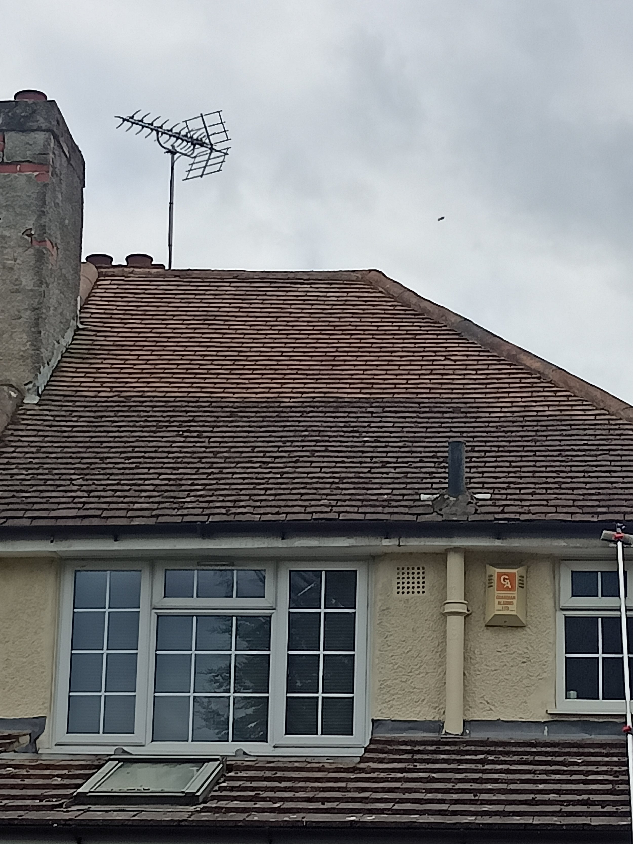 The top part of a building roof with a TV antenna and chimney against a cloudy sky, and a lower part showing large windows and a roof window.