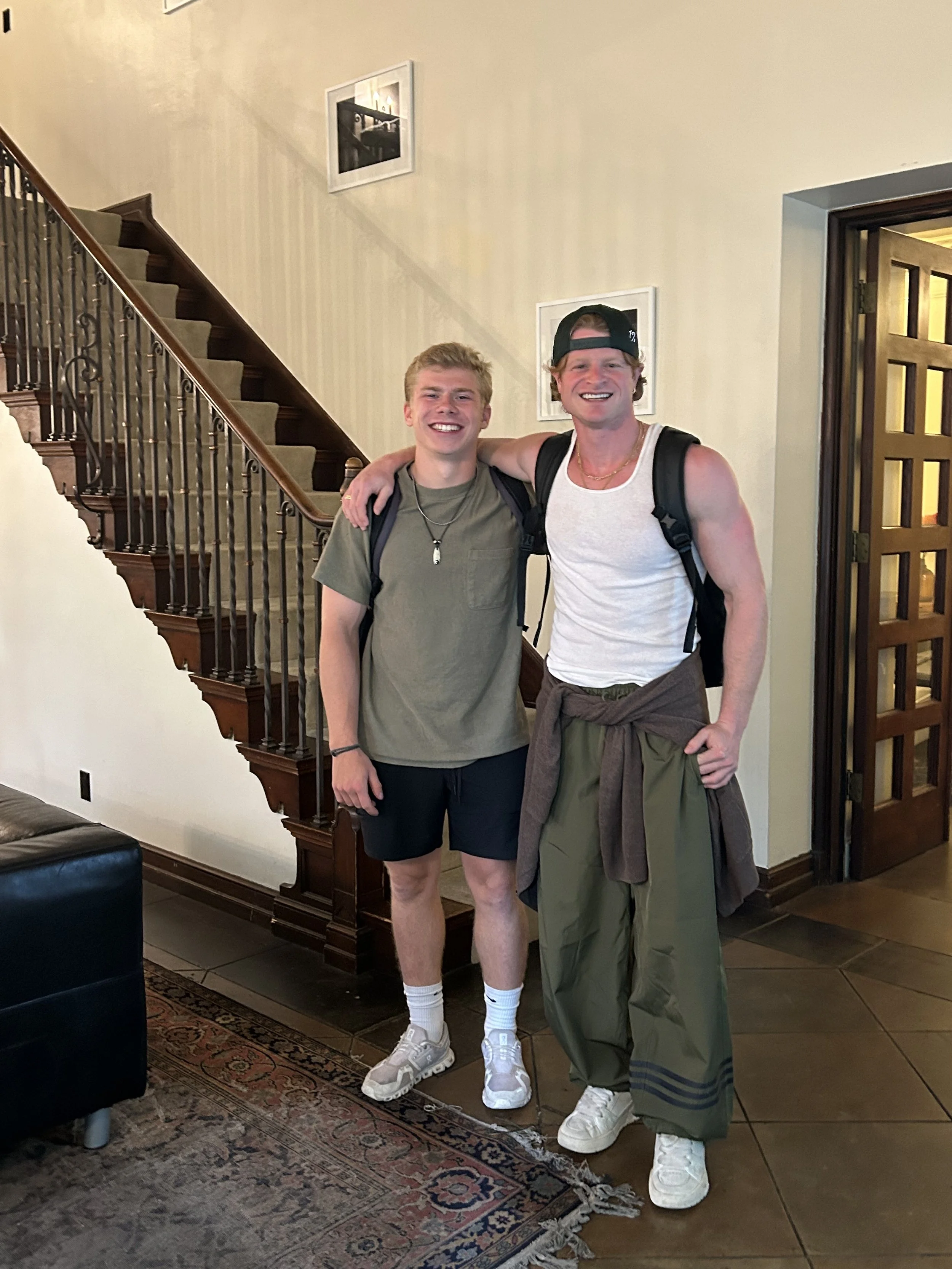 Two men standing inside a house, smiling at the camera, with one man's arm around the other's shoulder. They are casually dressed, wearing backpacks and athletic shoes.