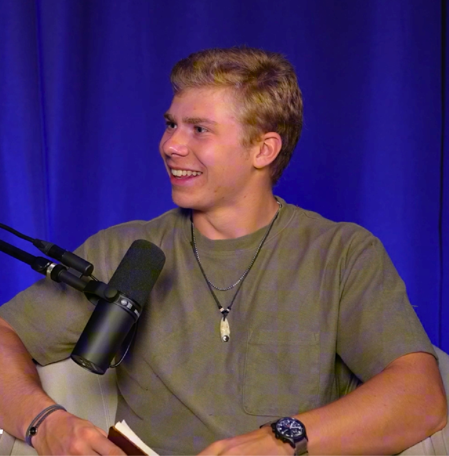 A young man with short blond hair smiling during a podcast recording, sitting in front of a professional microphone, wearing a green t-shirt, layered necklaces, and a wristwatch, with a blue curtain background.